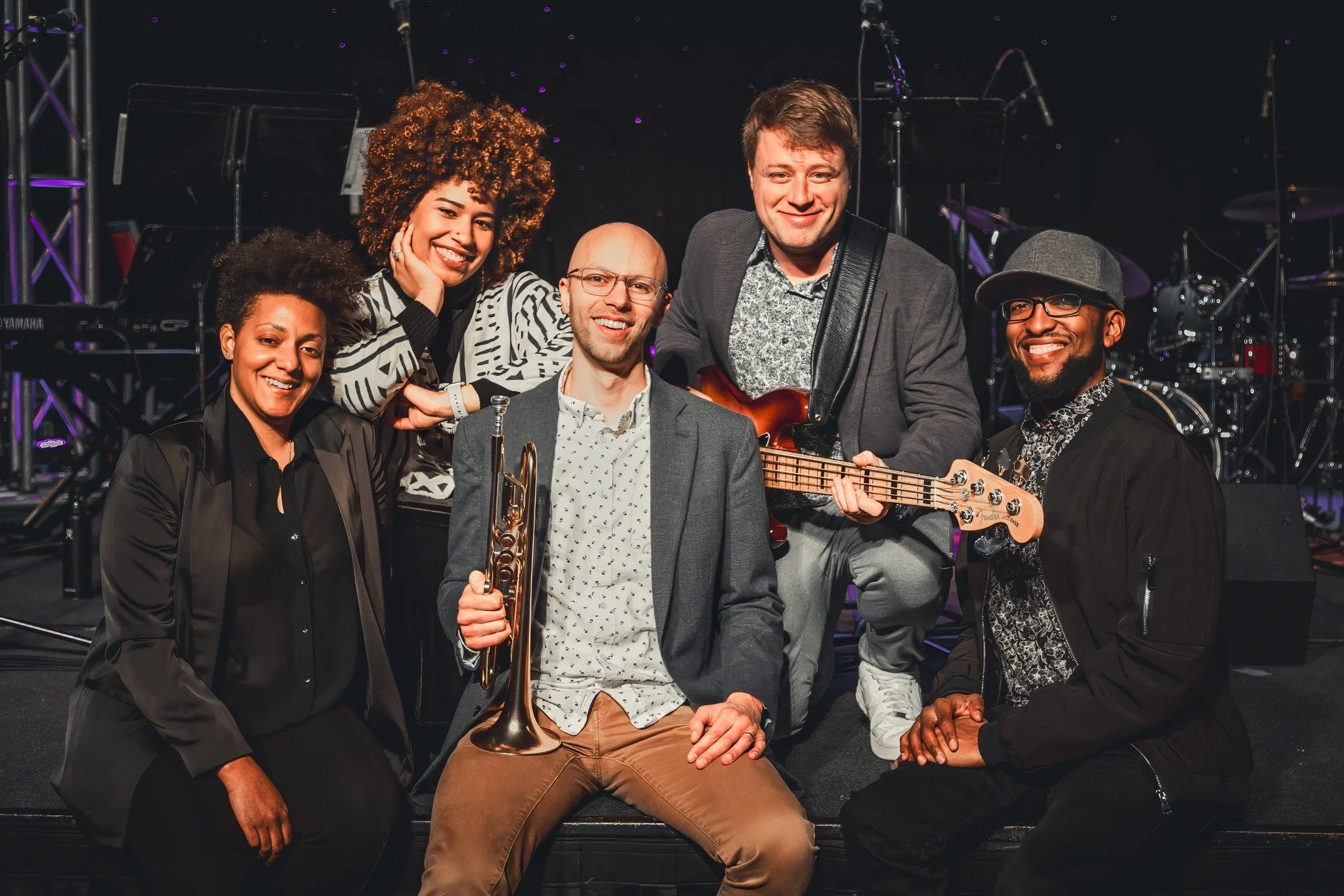 Group of six musicians smiling on stage with musical instruments, including a trumpet and bass guitar, against a dark background with stage equipment.