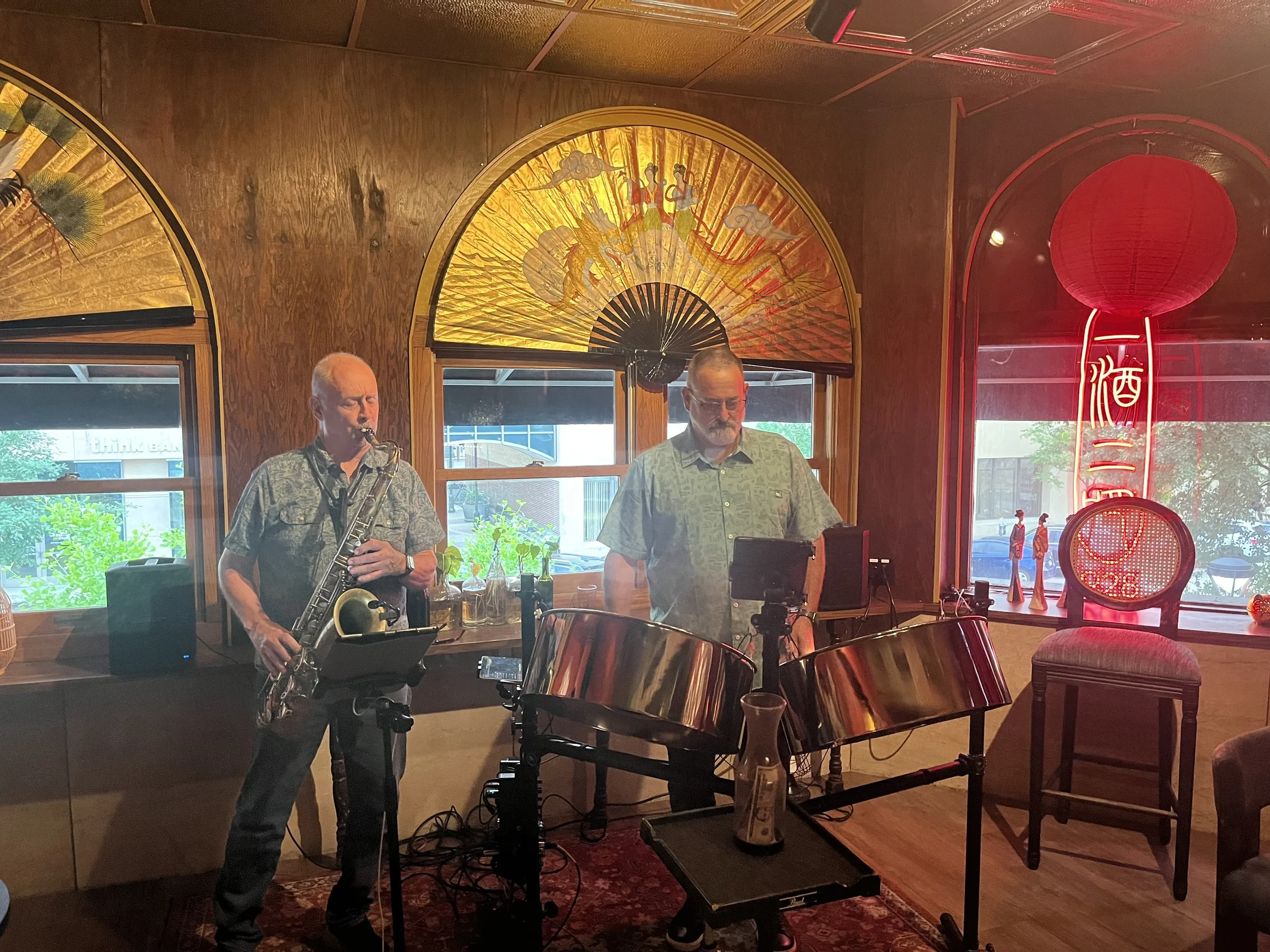 Two musicians performing in a cozy Asian-themed restaurant with wooden walls, decorative fans and red lanterns, one playing a saxophone and the other operating a keyboard.