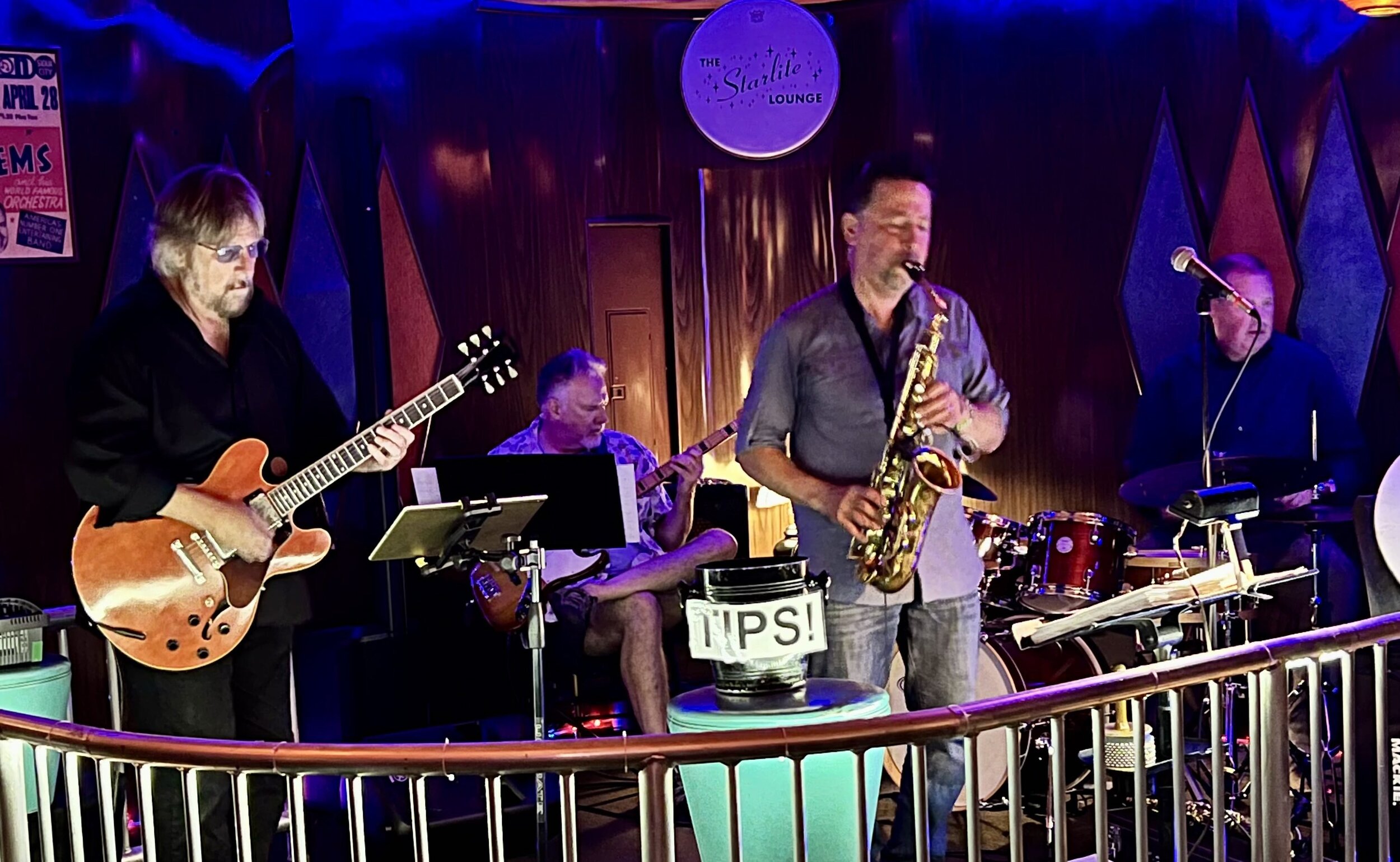 A jazz band performing live at The Starlite Lounge with four musicians playing guitar, saxophone, drums, and keyboard, on a wooden stage with a purple and red background.