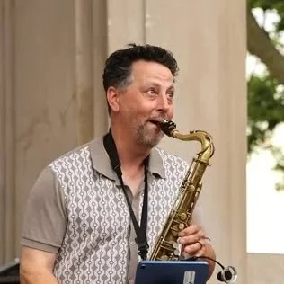 Man playing a saxophone outdoors near a stone building with trees in the background