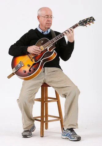 An older man sitting on a wooden stool playing an electric guitar against a plain white background.