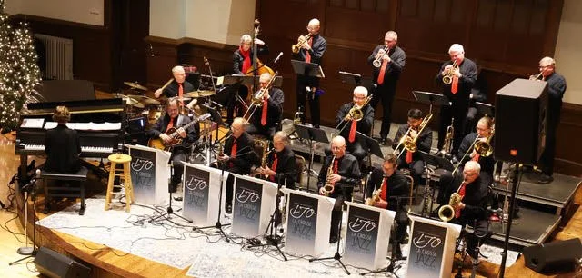 A jazz band performing on stage with a pianist, colored musicians, and a large group of wind instrument players, in a concert hall decorated with a Christmas tree.