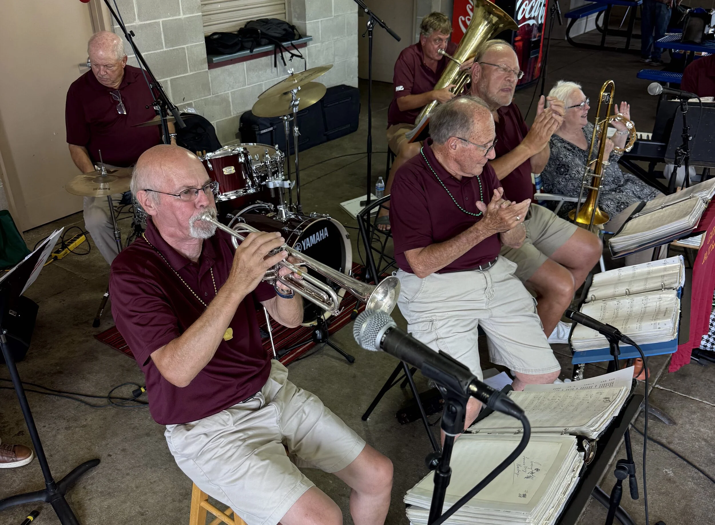 Group of senior musicians performing, including a man playing trumpet, a woman with a saxophone, and other band members playing various instruments, seated in a room with music stands and a microphone.