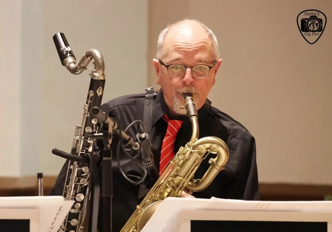 An older man wearing glasses, a black shirt, and a red tie is playing a saxophone. There are music stands with sheet music in front of him, and he is in a room with beige walls.