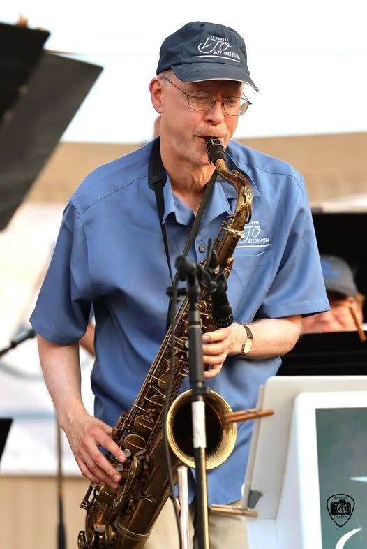 A man playing a saxophone at an outdoor music event, wearing glasses, a blue short-sleeved shirt, and a cap.