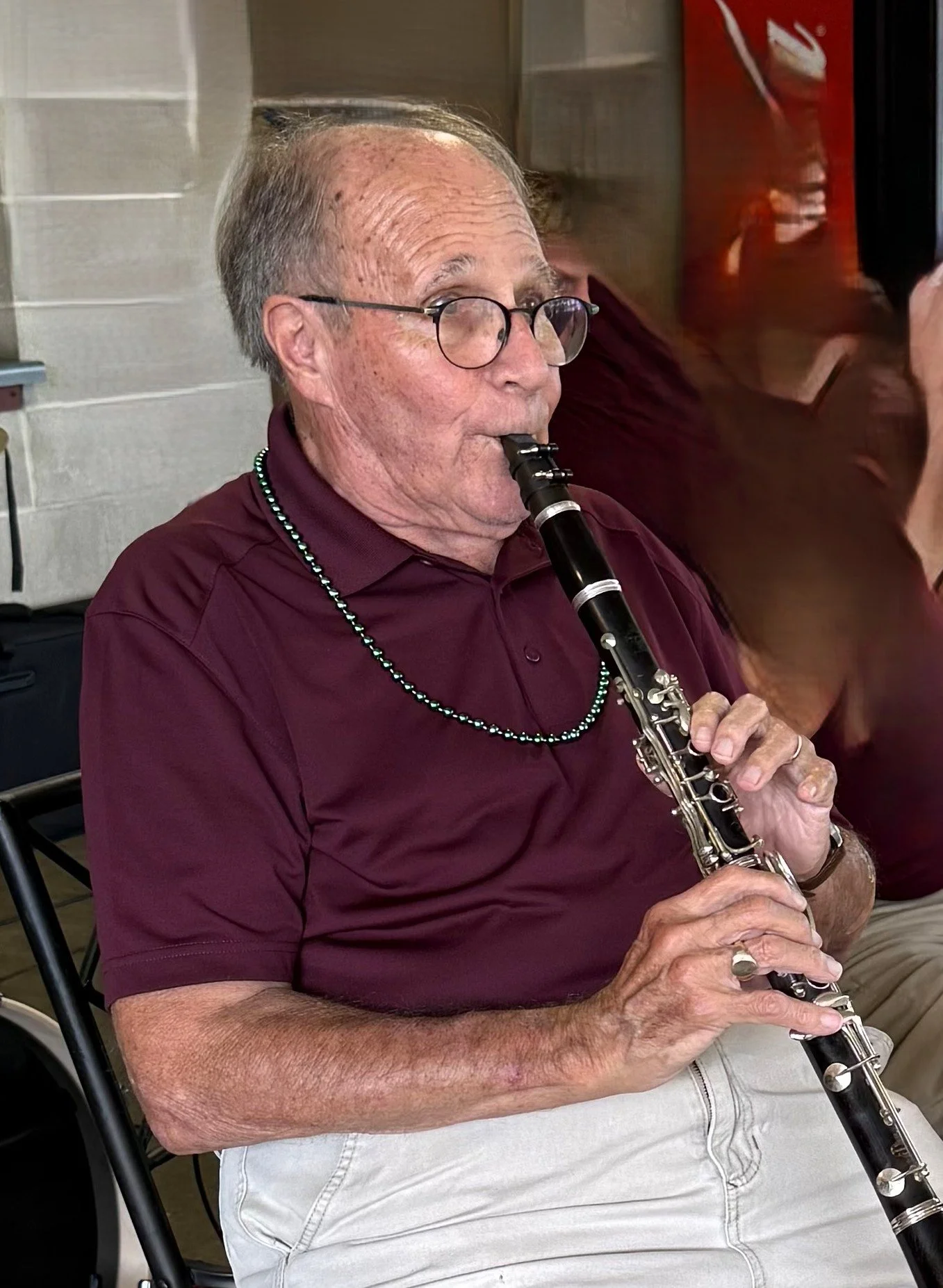An elderly man wearing glasses, a maroon polo shirt, a beaded necklace, and a wedding ring is playing a black clarinet. He appears to be seated indoors, and there is a woman in the background sitting next to him, partially visible.
