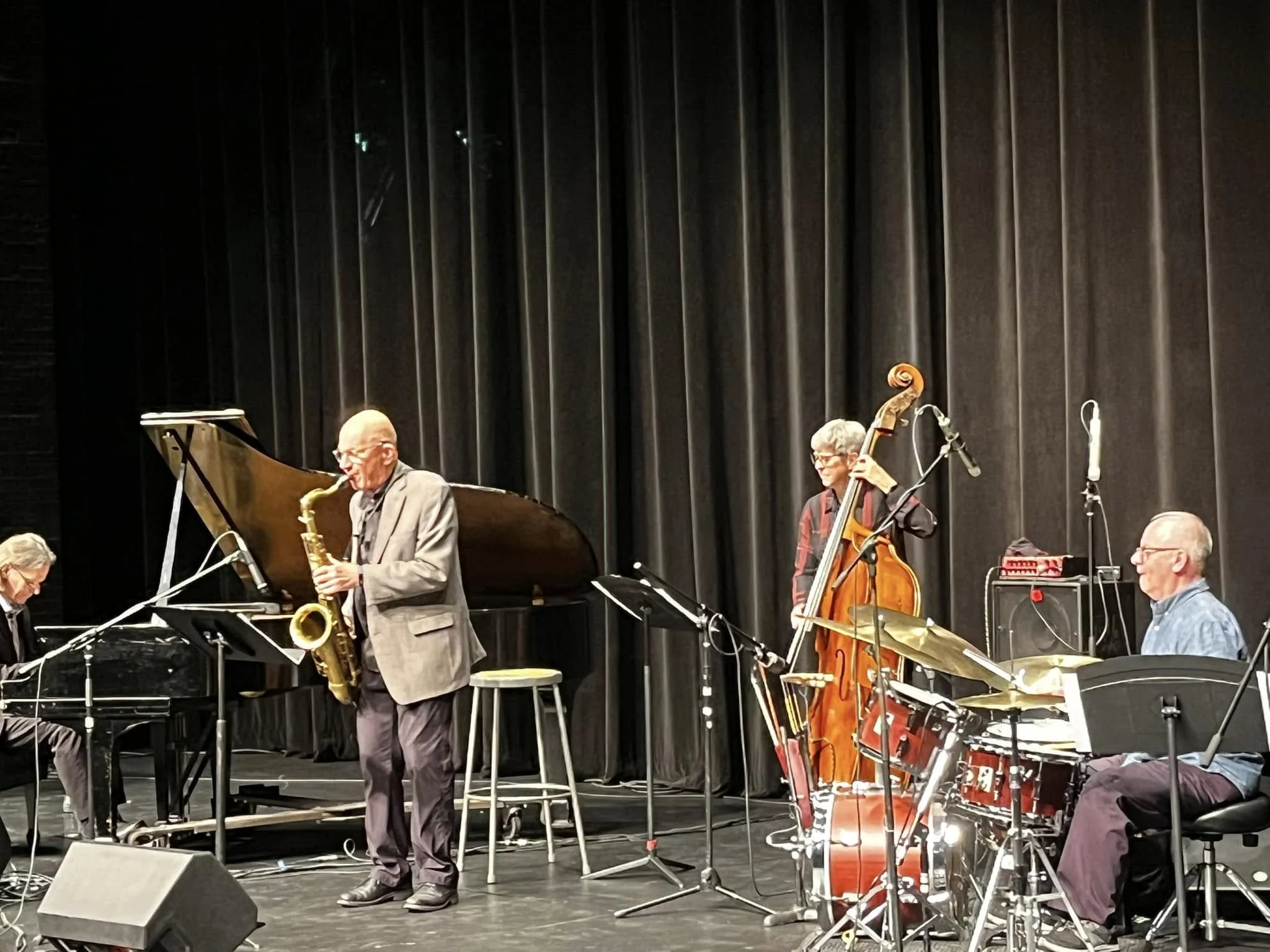 Four jazz musicians perform on stage with a grand piano, a saxophone, a double bass, and a drum set, in front of black curtains.
