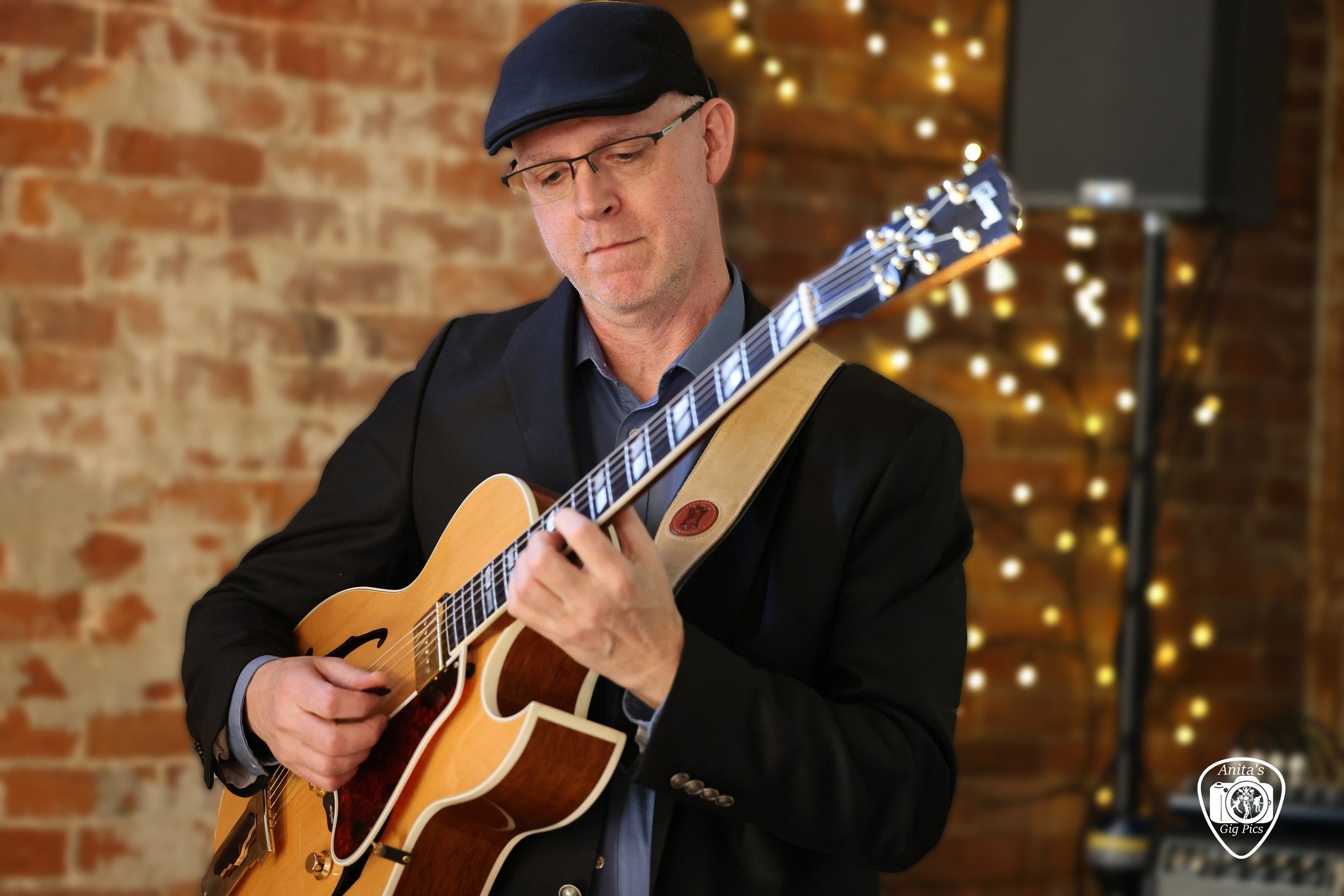 A man wearing glasses and a flat cap playing an acoustic guitar against a brick wall with string lights and a speaker in the background.