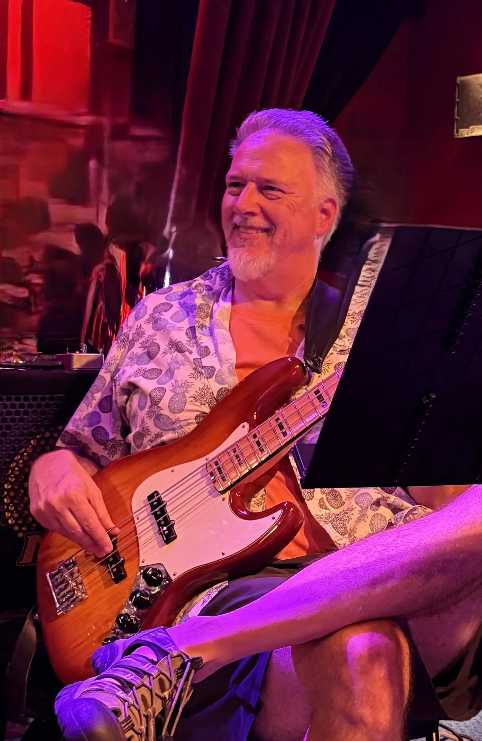 A smiling man with gray hair and facial hair playing an electric bass guitar while sitting on a chair in a dimly lit room with purple and red lighting, wearing a short-sleeved shirt with a pineapple pattern.