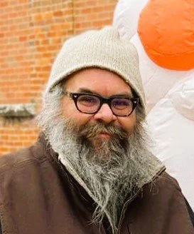 A man with glasses, a gray beard, and a beige beanie smiling outdoors near a large inflatable snowman's nose.