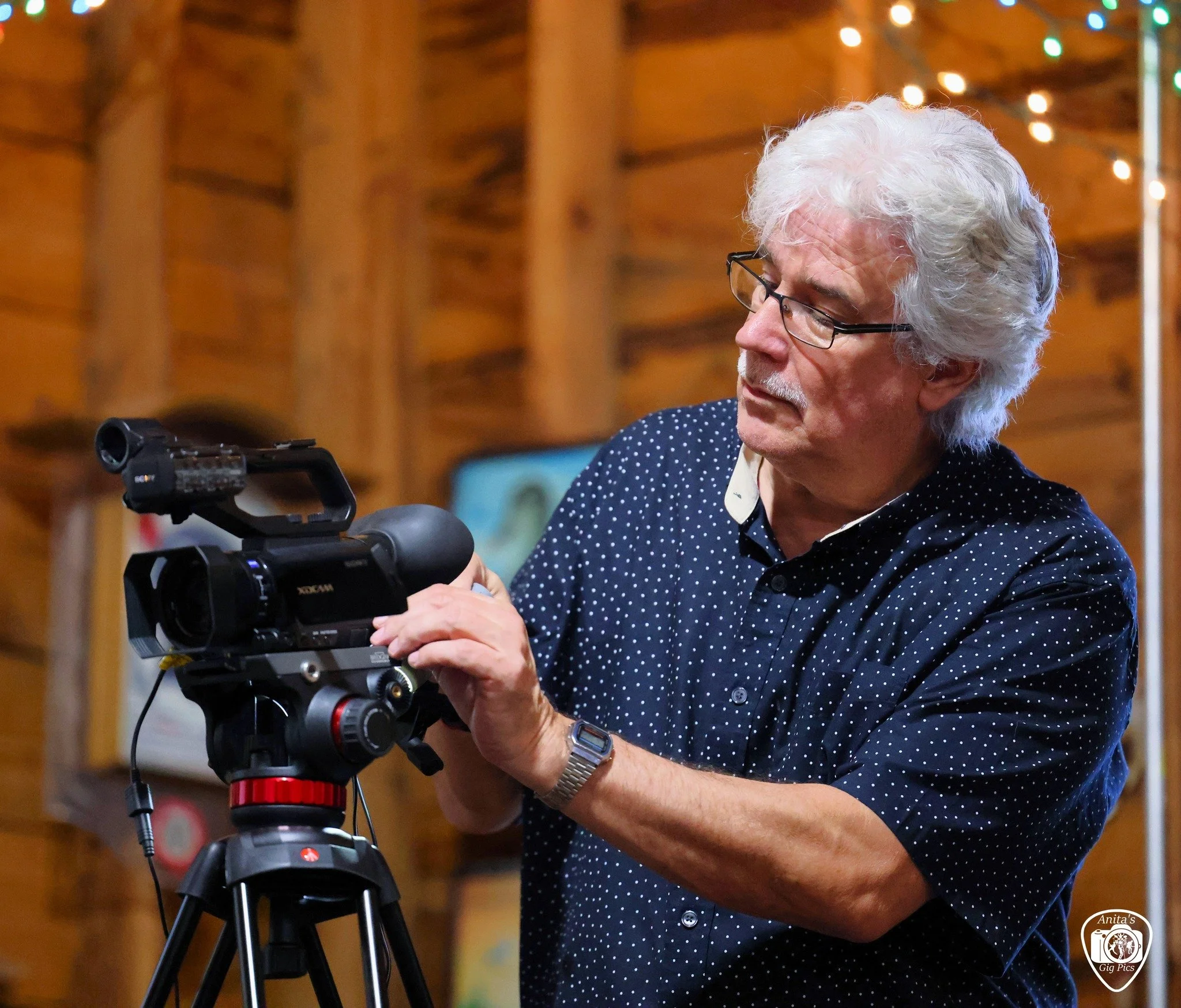 An elderly man with white hair and glasses adjusting a professional video camera on a tripod indoors with wooden walls and string lights.