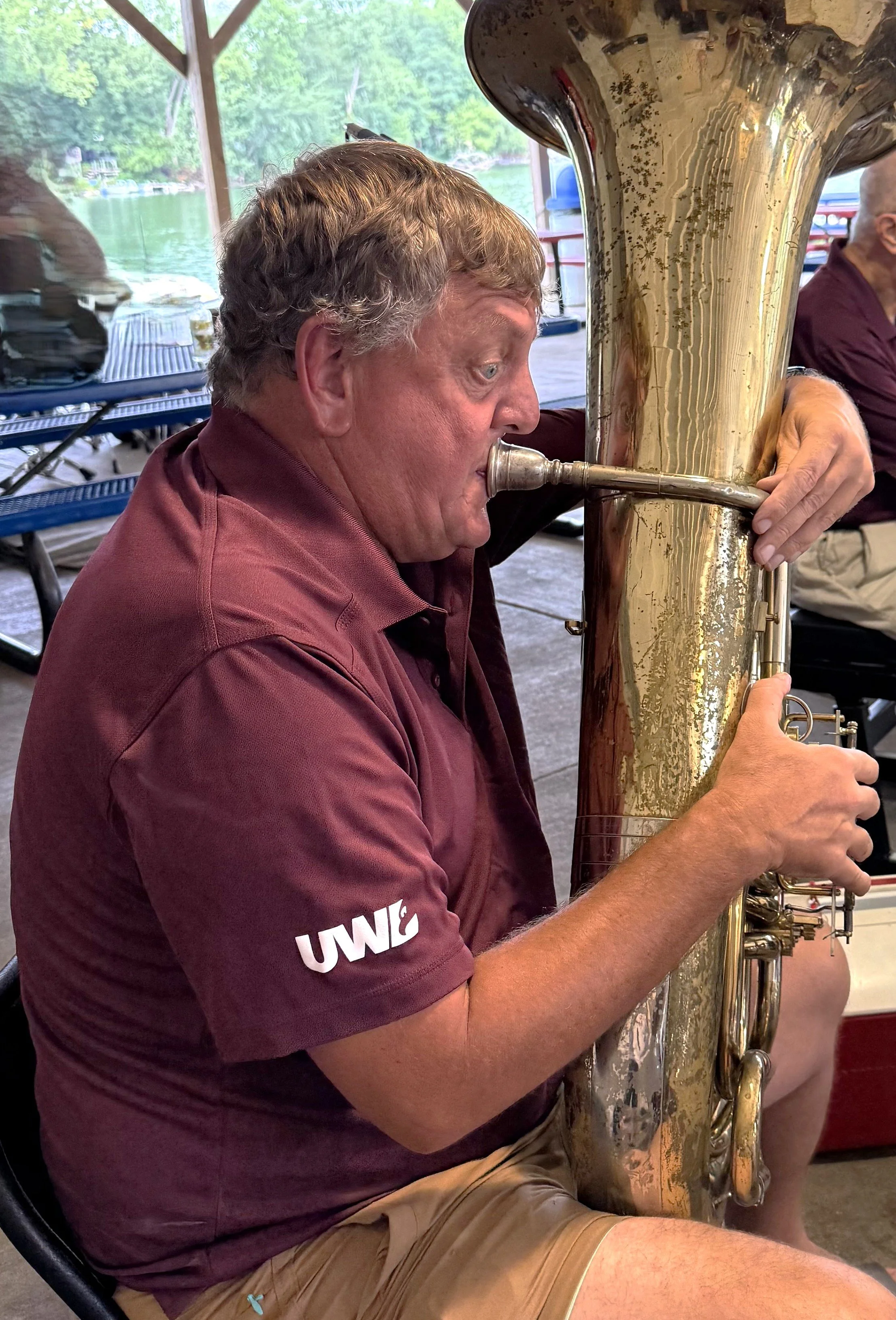 A man playing a large brass tuba at a lakeside or riverside outdoor setting.