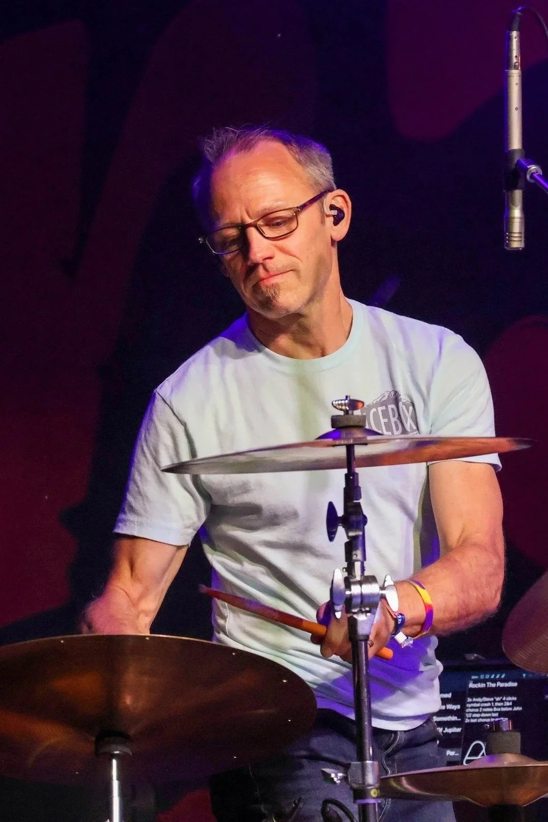A middle-aged man with glasses and a goatee playing drums on stage, wearing a light gray T-shirt, with a microphone stand nearby and a dark background.