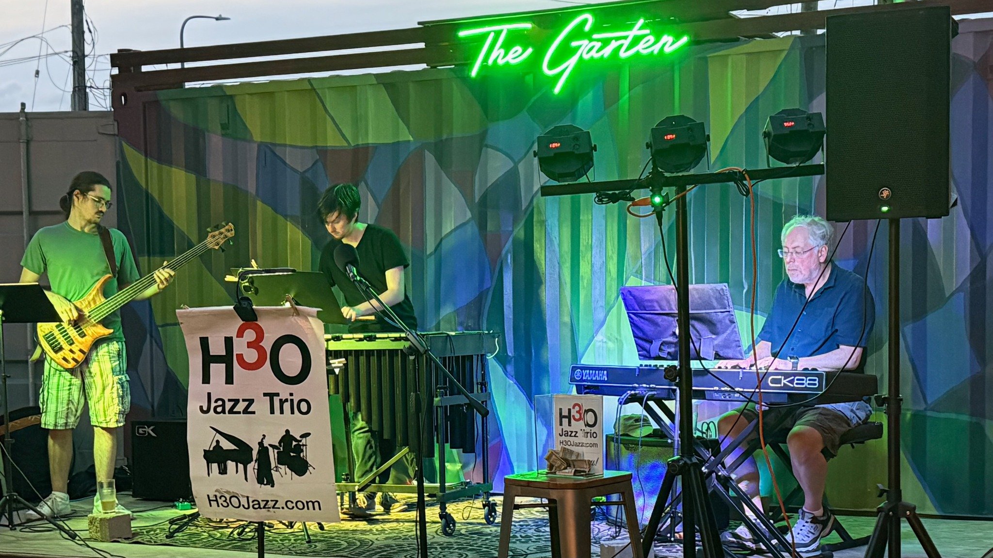 A jazz trio performing on a stage with a neon sign that reads 'The Garden.' The band consists of a bass player on the left, a vibraphone player in the middle, and a keyboardist on the right. There is a banner in front of the vibraphone with the text 