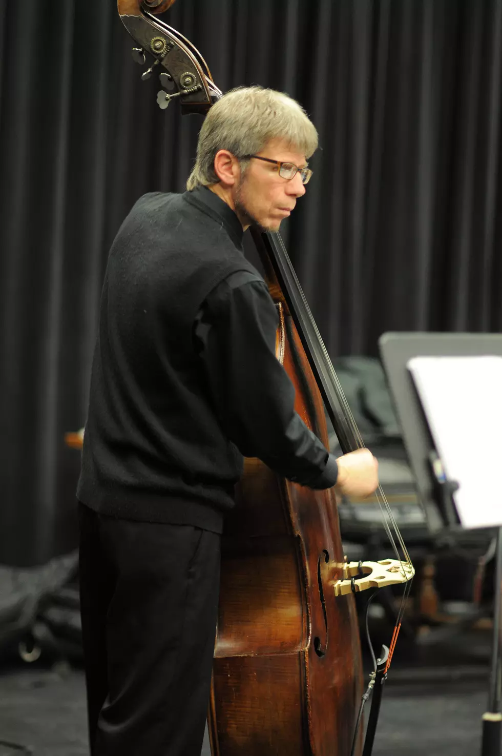 A man with glasses and gray hair is playing a double bass in a music rehearsal or performance setting, with a black curtain in the background.