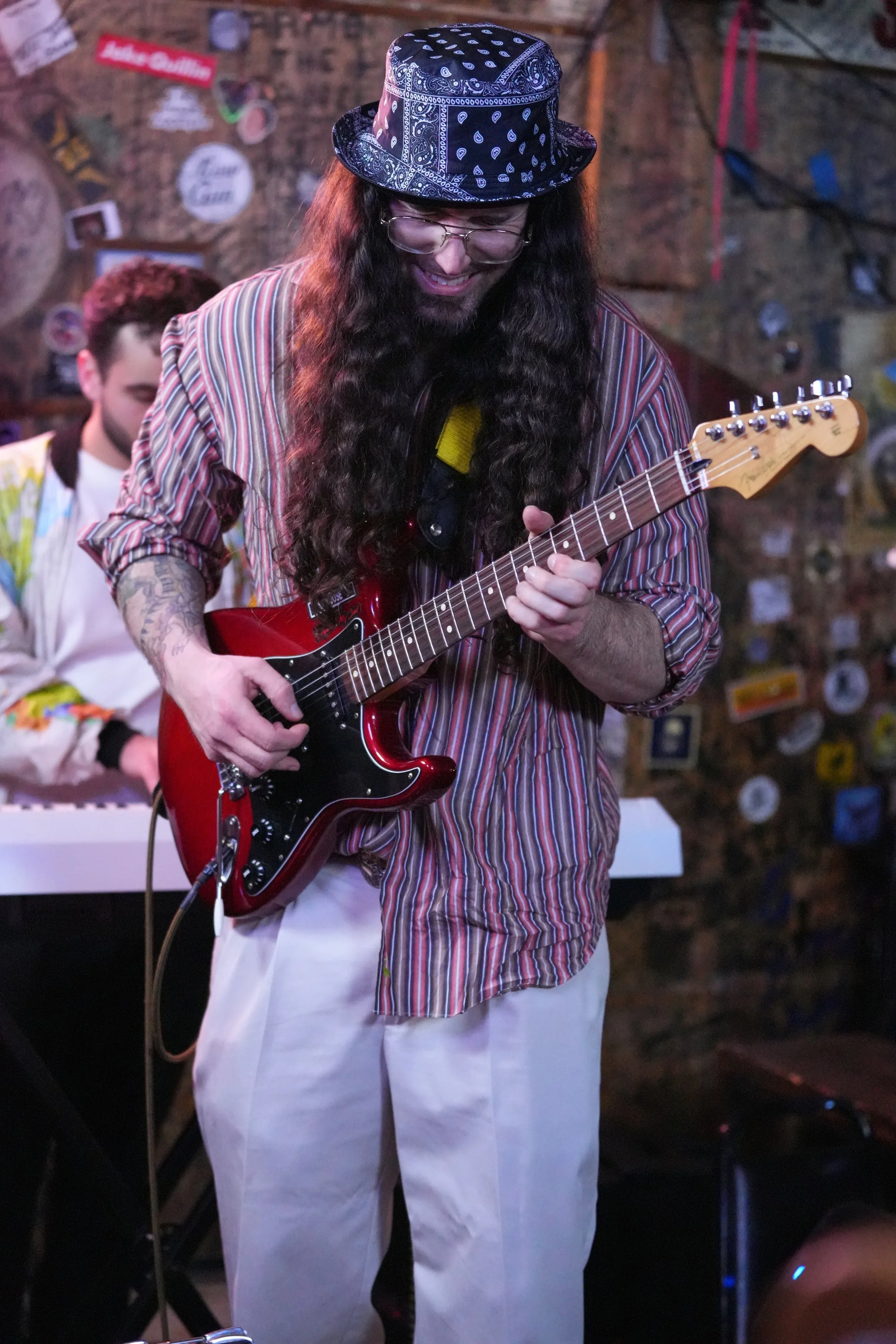 A man with long curly hair, glasses, and a bandana hat playing a red electric guitar in a dimly lit, decorated venue. Carson Lewis Music