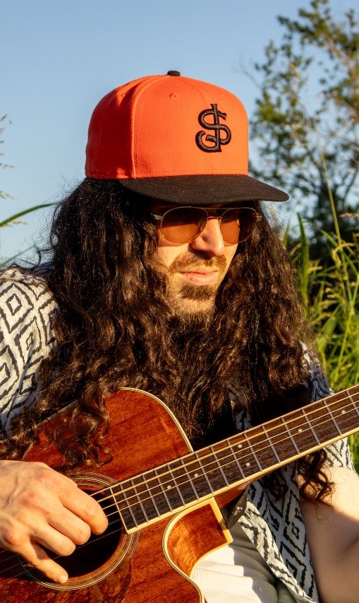 A man with long curly hair and a beard playing an acoustic guitar outdoors, wearing an orange and black cap, sunglasses, and a patterned shirt. Carson Lewis Music