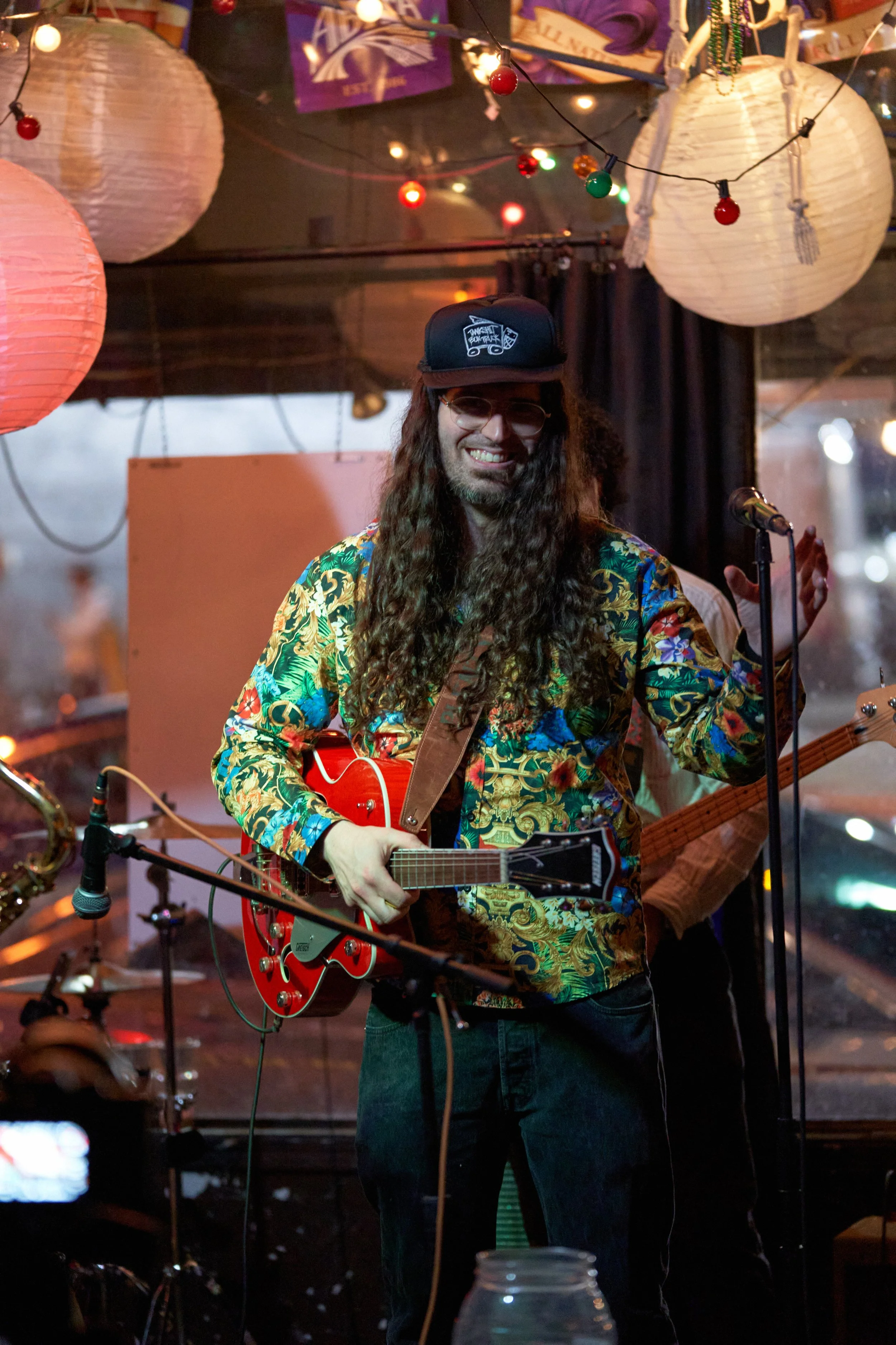A musician with long curly hair, wearing glasses, a colorful floral shirt, and a black cap, playing a red electric guitar and smiling on stage at a lively, decorated venue with paper lanterns and string lights. Carson Lewis. Deep Ellum