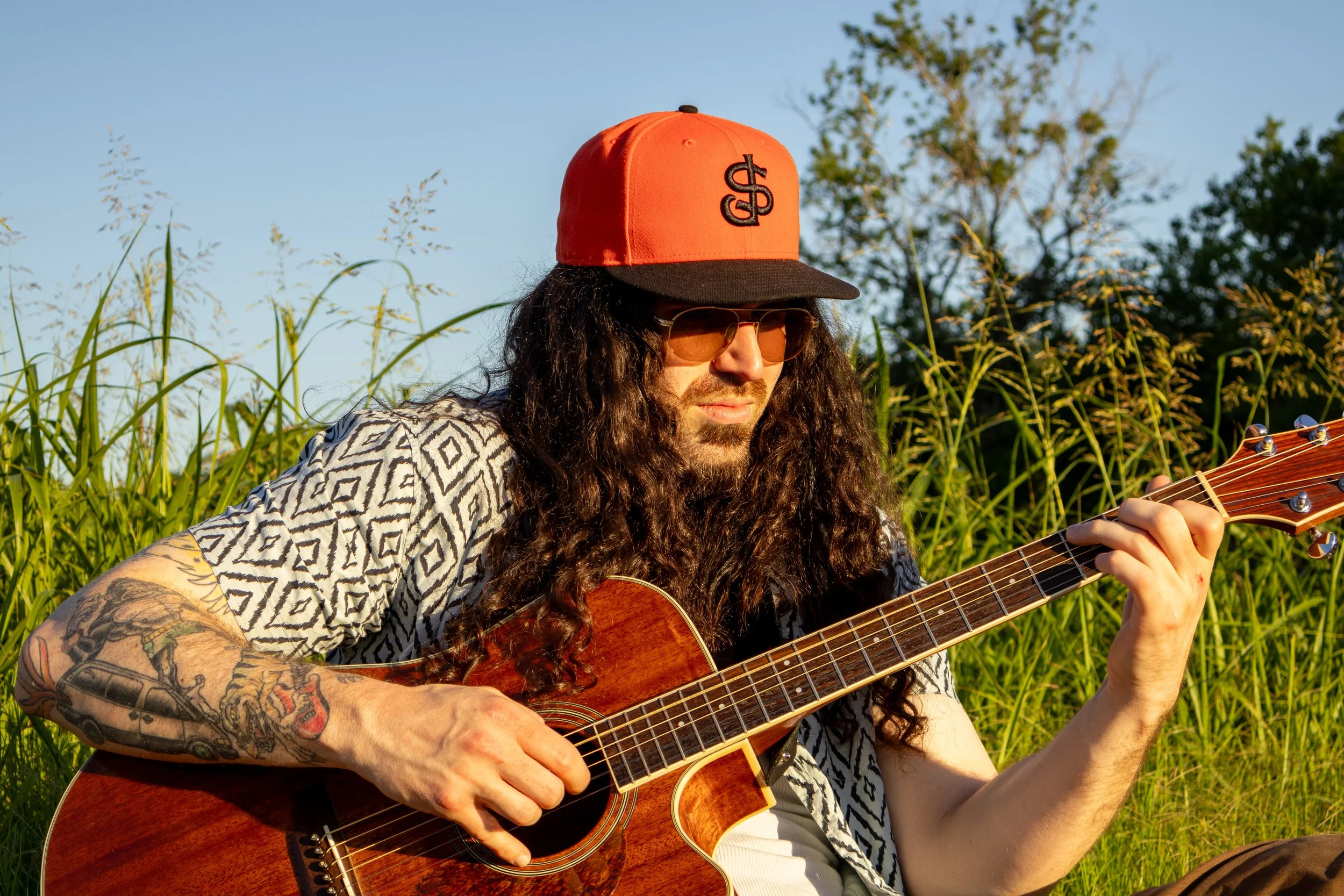 A man with long curly hair, wearing sunglasses and a red and black baseball cap, playing an acoustic guitar in a field of tall grass with trees in the background, during daylight. Carson Lewis