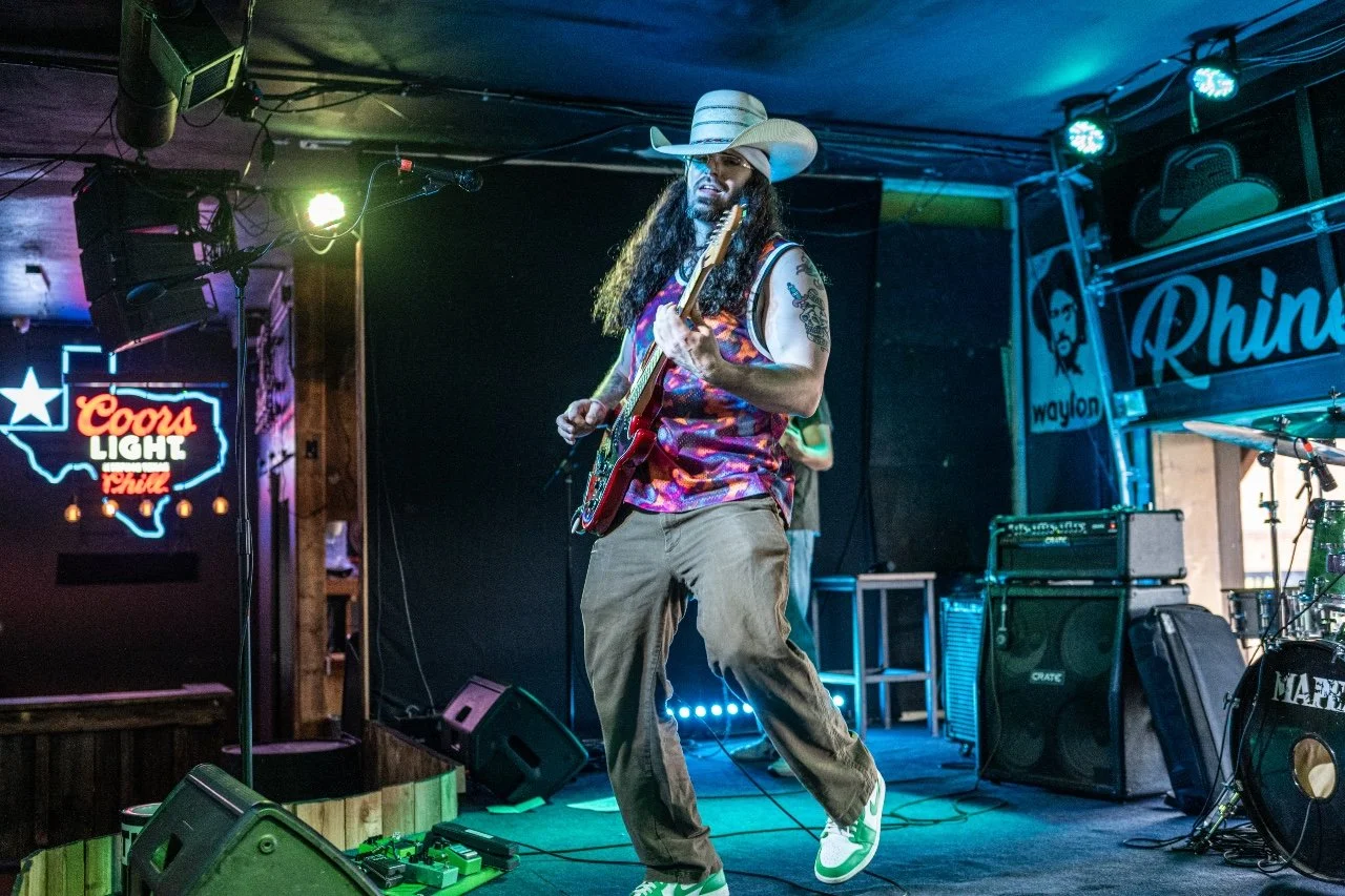 A man with long hair and tattoos, wearing a cowboy hat, colorful tank top, and brown pants, playing an electric guitar on stage at a bar or music venue with neon signs and musical equipment in the background. Carson Lewis Music