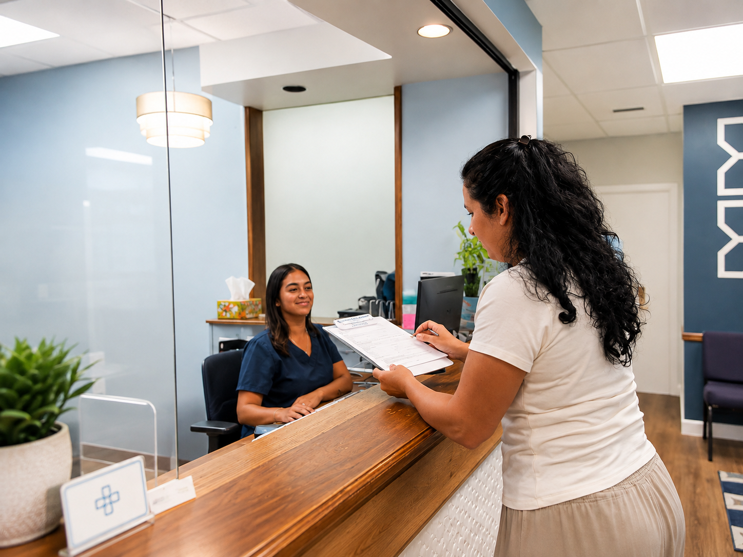 A woman with curly black hair in a white shirt checks a clipboard while talking to a female receptionist with straight brown hair in a navy uniform at a reception desk with plants and a tissue box.
