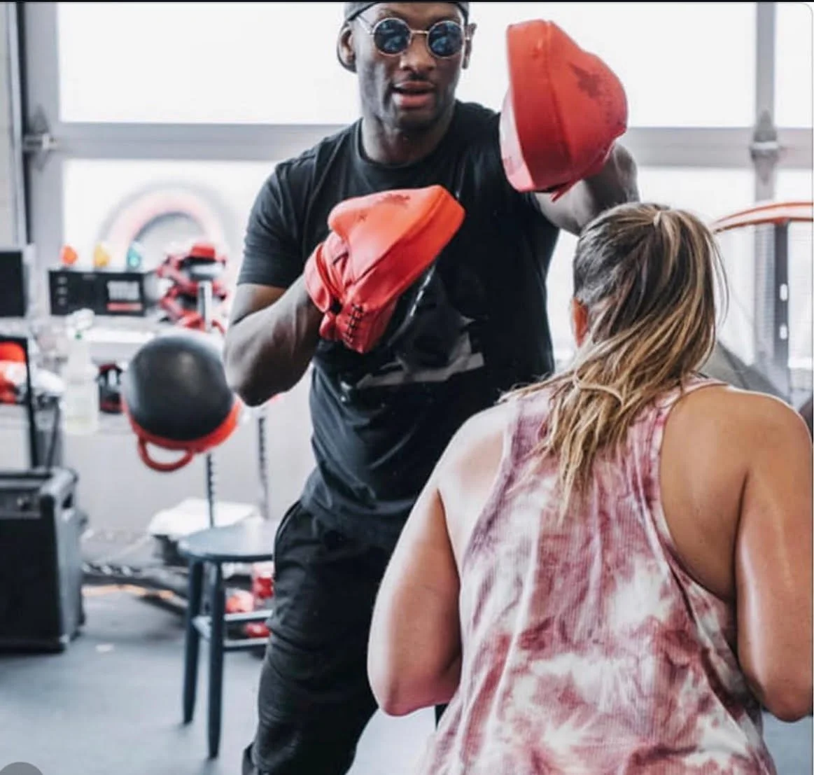 A man wearing sunglasses and boxing gloves is punching a woman with long hair in a gym. The woman is wearing a pink tank top and is facing away from the camera. The gym has various equipment and weights in the background.
