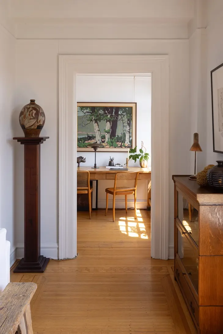 View through a doorway into a sunlit room with a wooden desk, two matching chairs, a painting of trees on the wall, a potted plant, and a small sculpture.