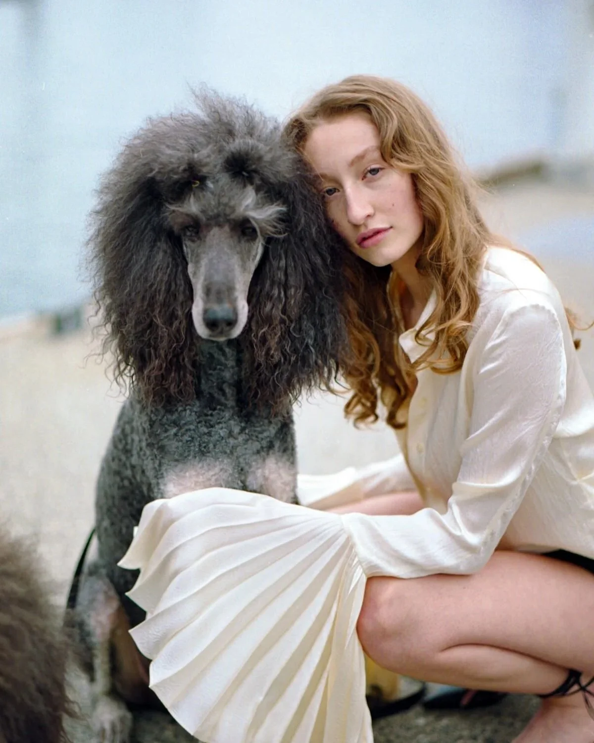 A woman with curly red hair and a white dress sitting on the ground next to a large black poodle with curly fur, both resting their heads together.