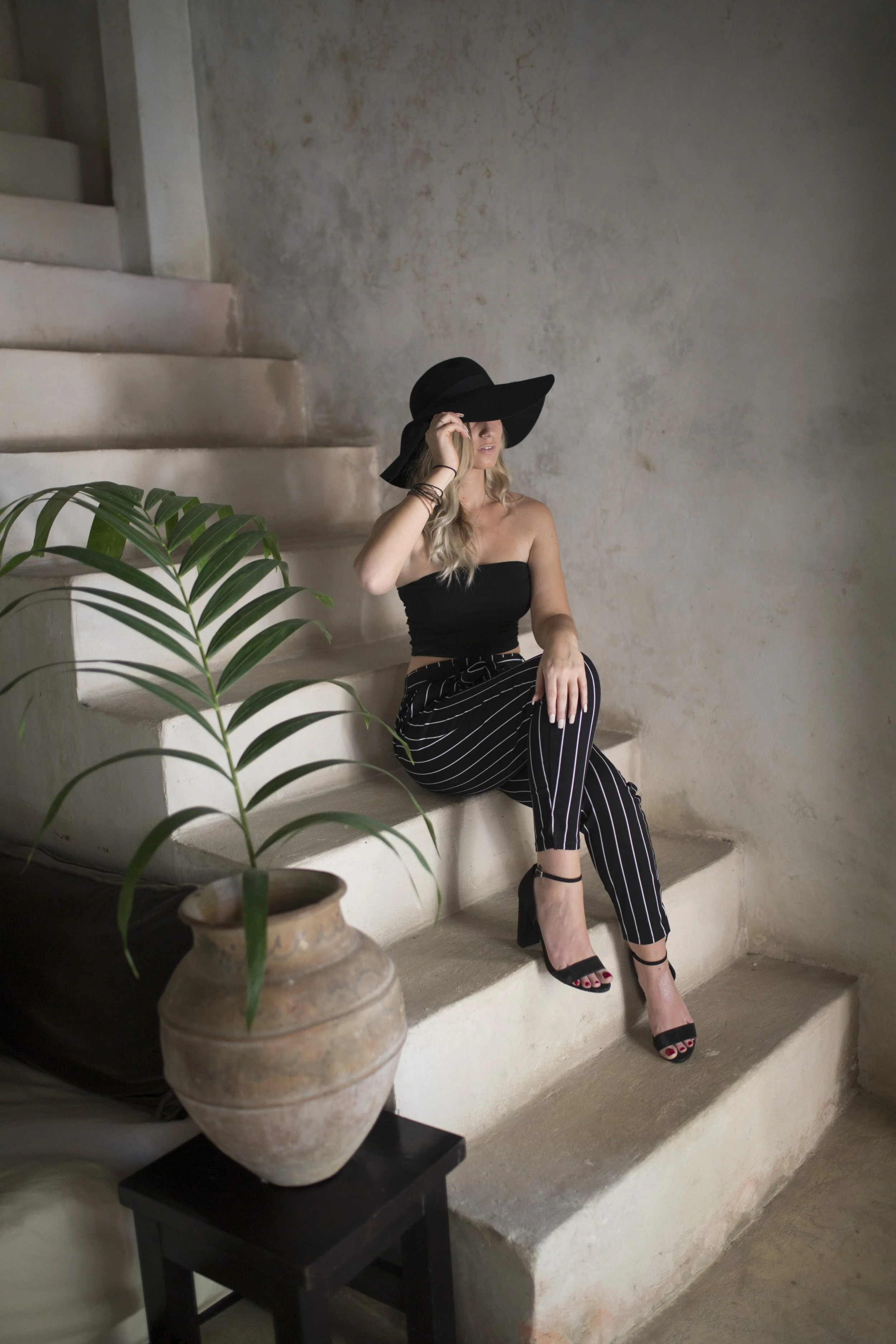 A woman sitting on stairs wearing a black strapless top, black and white striped pants, black high-heeled shoes, and a wide-brimmed black hat, with a large potted plant nearby.