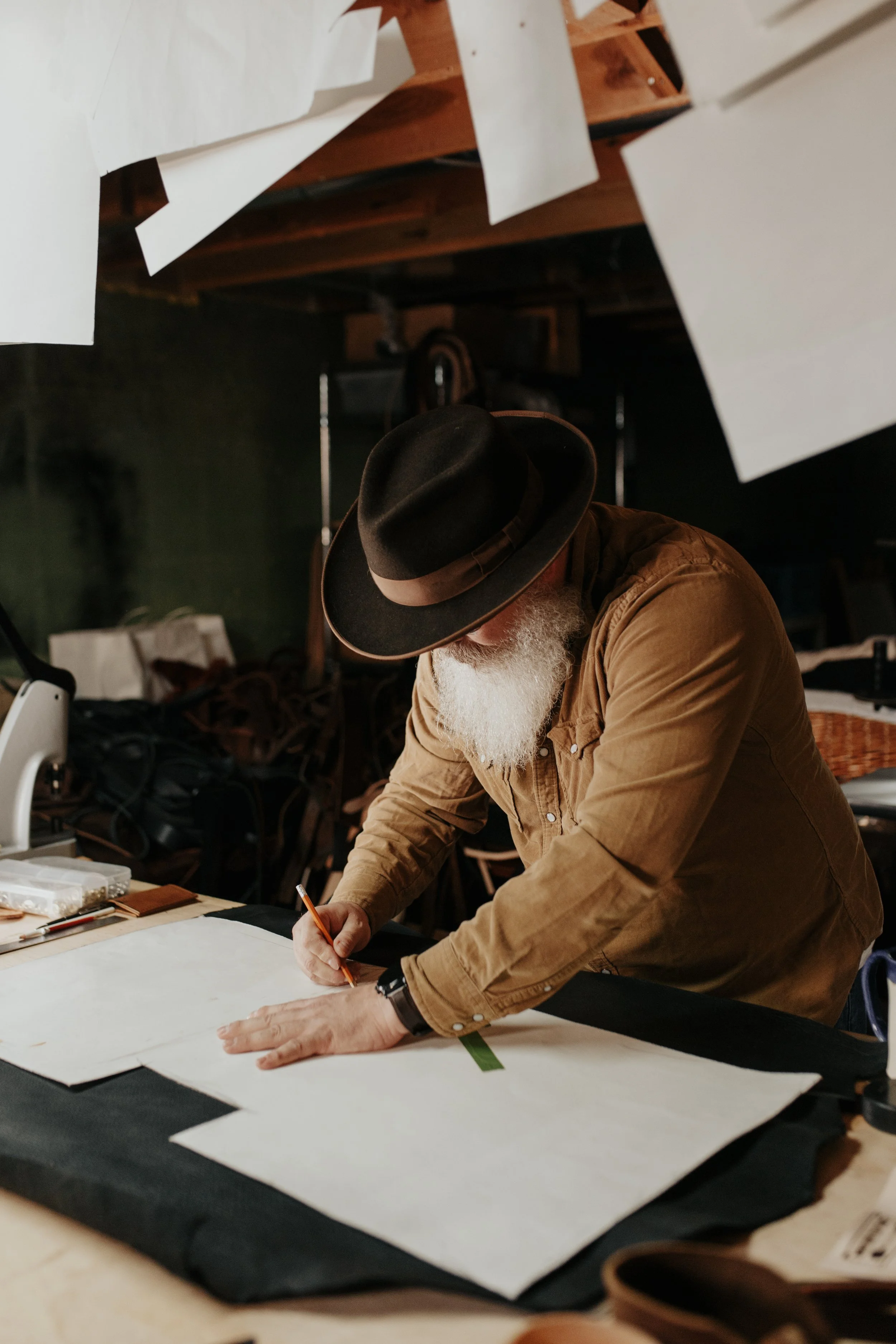 An older man with a white beard wearing a brown shirt and black hat working on a large piece of fabric or paper on a worktable, in a workshop setting.