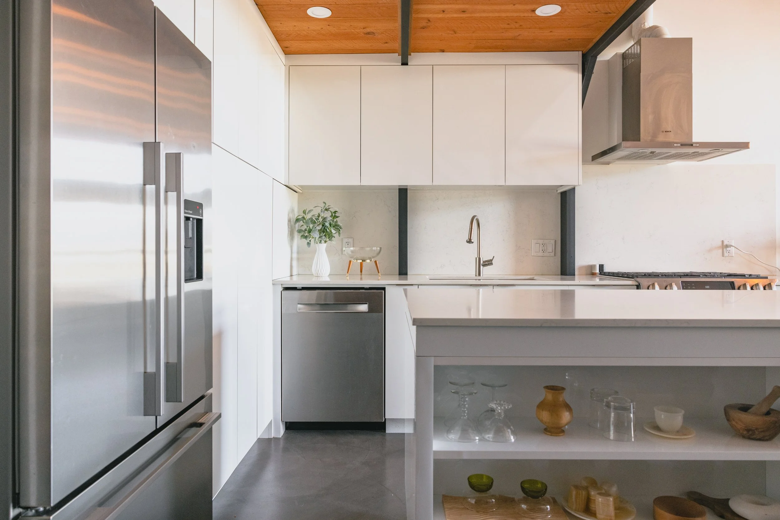 Modern kitchen with stainless steel appliances, white cabinetry, and a wooden ceiling. Features a plant in a white vase, glassware, and a kitchen island with open shelves.