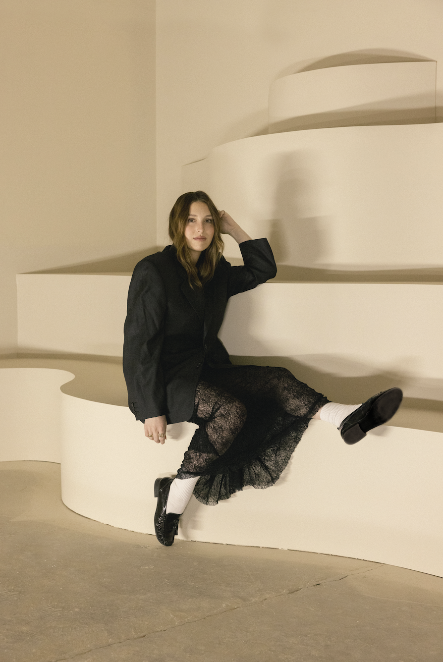 A woman sitting on a white curved platform, wearing a black blazer, a black lace skirt, white socks, and black shoes, with a neutral expression, in a minimalist indoor setting with soft lighting.