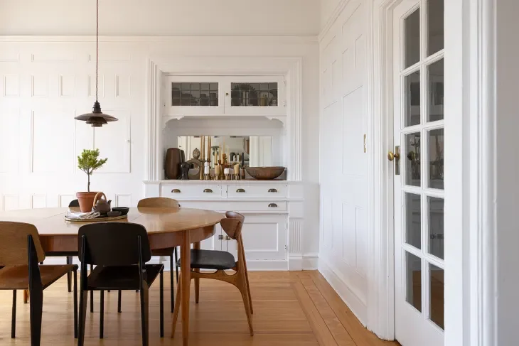 Dining room with a wooden table and black and brown chairs, white built-in cabinetry with a mirror, decorative items, and a small potted plant, with hardwood flooring.