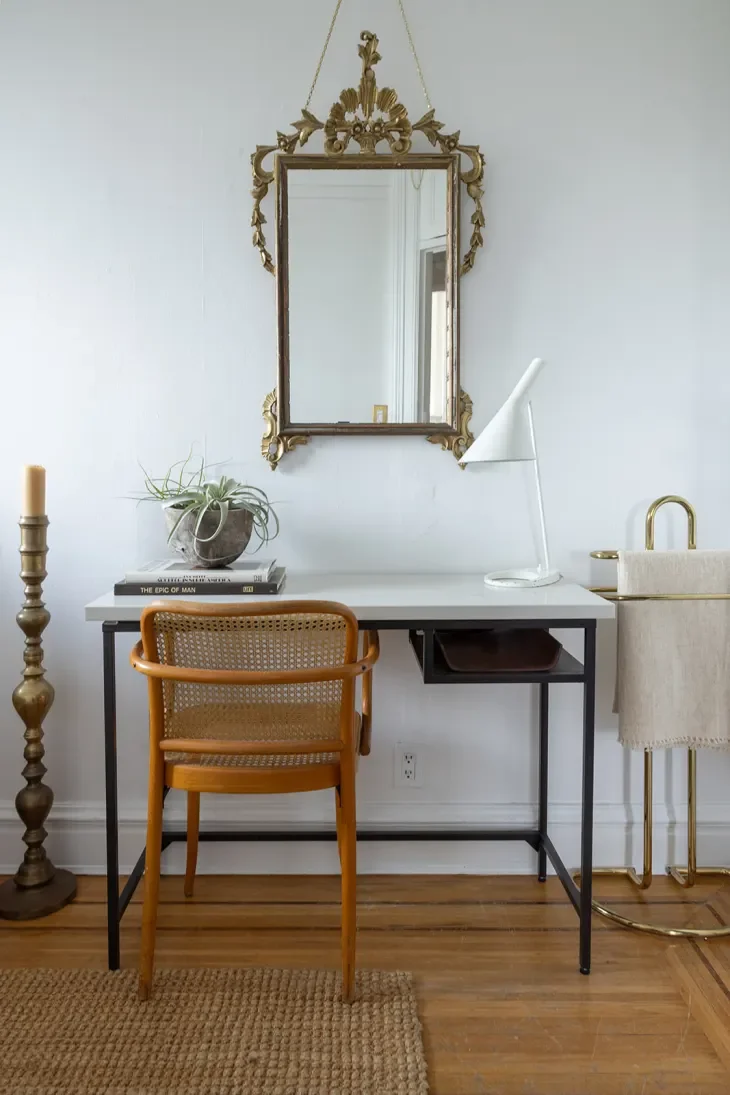 A white desk with black legs against a white wall, framed mirror with ornate gold frame hanging above, a tan chair with cane back, a white table lamp, a potted plant, and candles, with hardwood floor and a woven rug.