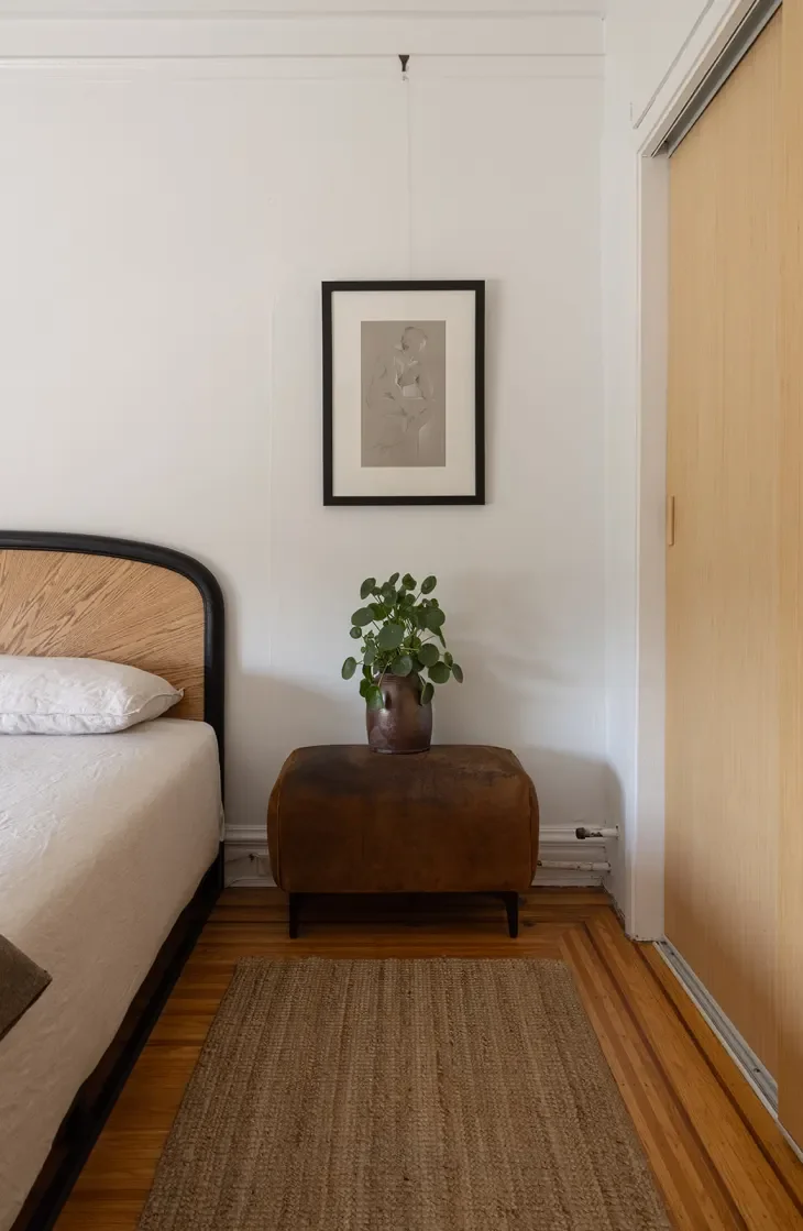 A bedroom corner with a wooden bed frame, a small wooden side table with a houseplant in a brown pot, a framed black-and-white art piece on the wall, and a beige rug on a wood floor. There is a closet door to the right.