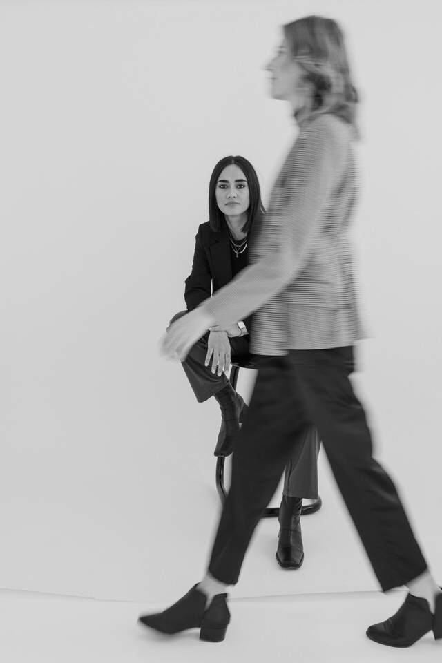 A woman sitting on a chair and looking directly at the camera, with another woman walking in front of her, blurred from motion, in a studio setting with a plain light background.