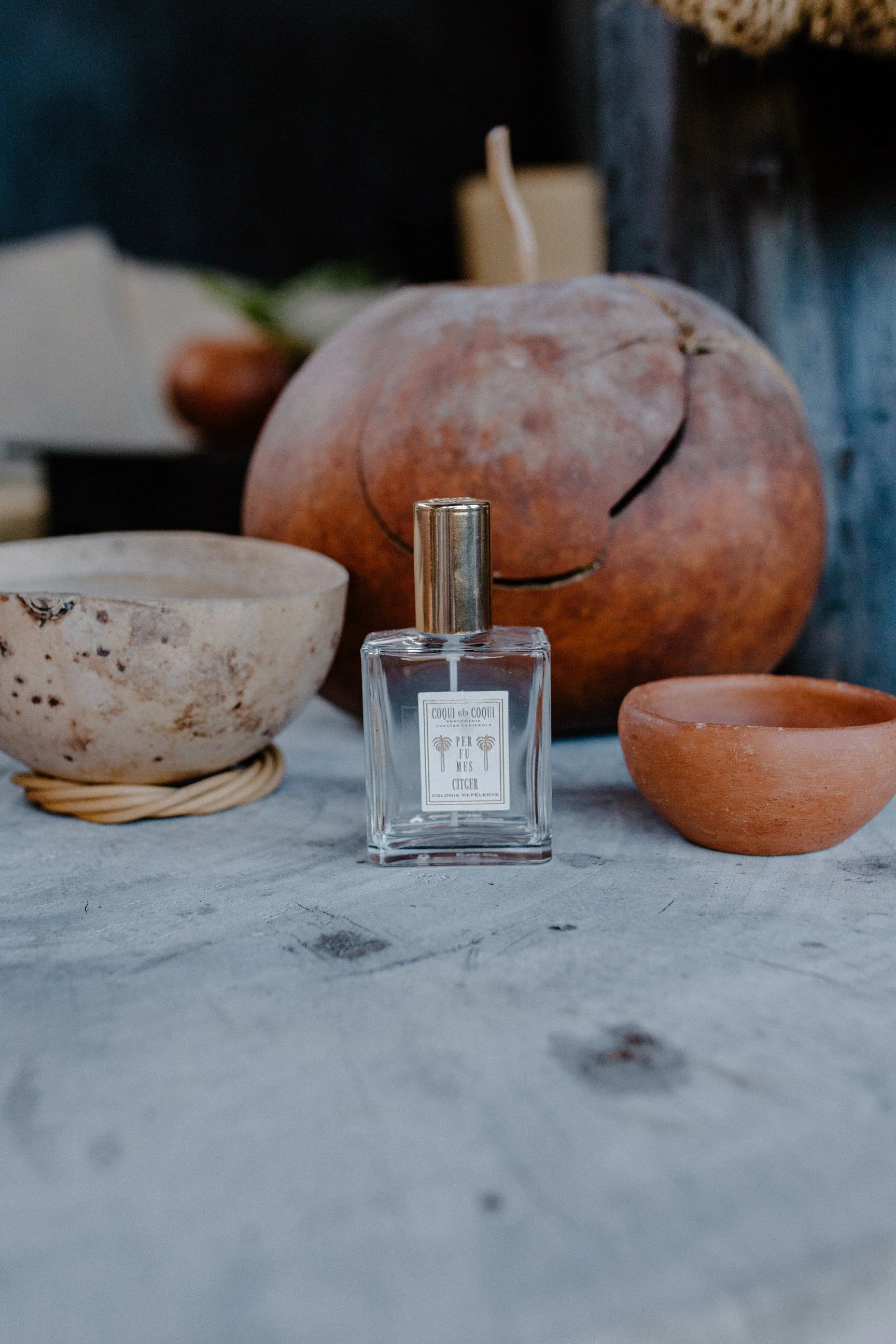 A glass bottle of perfume placed on a wooden surface, with ceramic bowls and a large, cracked pumpkin in the background.