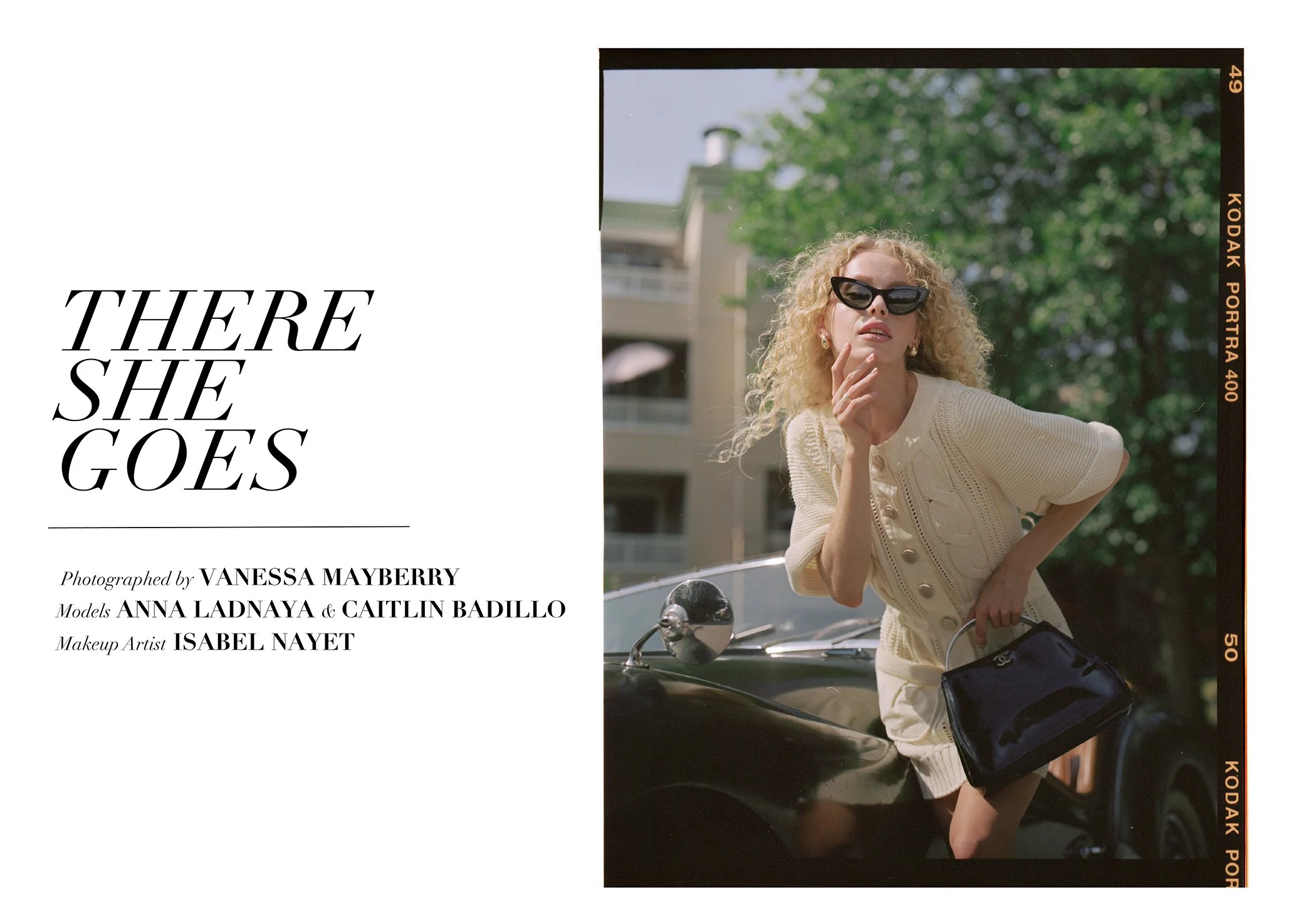 A woman with curly blonde hair wearing sunglasses, a cream-colored knit top, and holding a matching cream skirt and black handbag, leaning on a vintage black car, with a background of green trees and a multi-story building.