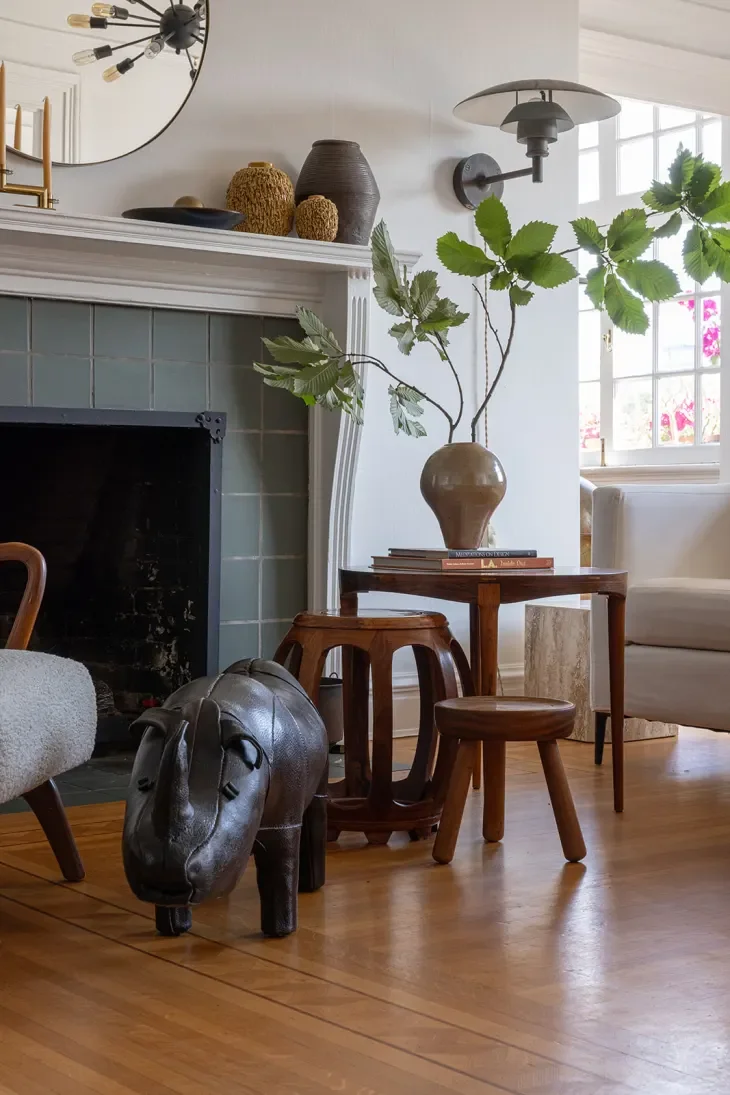 Living room with a black horse-shaped sculpture on the wooden floor, a small wooden table with a large vase and books, a white armchair, a fireplace with teal tiles, a large round wall mirror, and window with pink flowers outside.