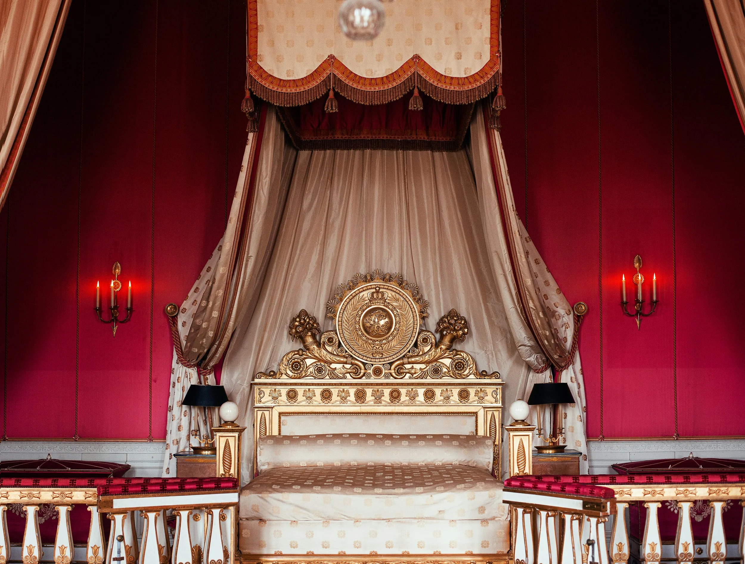A luxurious royal bed with ornate gold and cream detailing, rich silk curtains, and a deep red background. The bed has a high headboard with intricate carvings and is flanked by elegant side tables with lamps and candles.