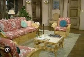 Living room straight out of the 1980s with pink floral sofas, yellow armchair, glass coffee table, and decor including a lamp and indoor plants.