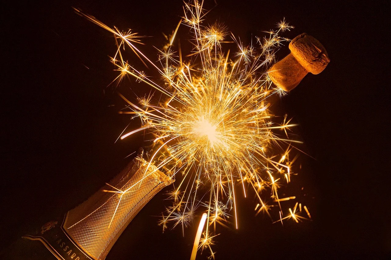 A bottle of champagne opening with a sparkler and cork flying out amid bright sparks in the dark.