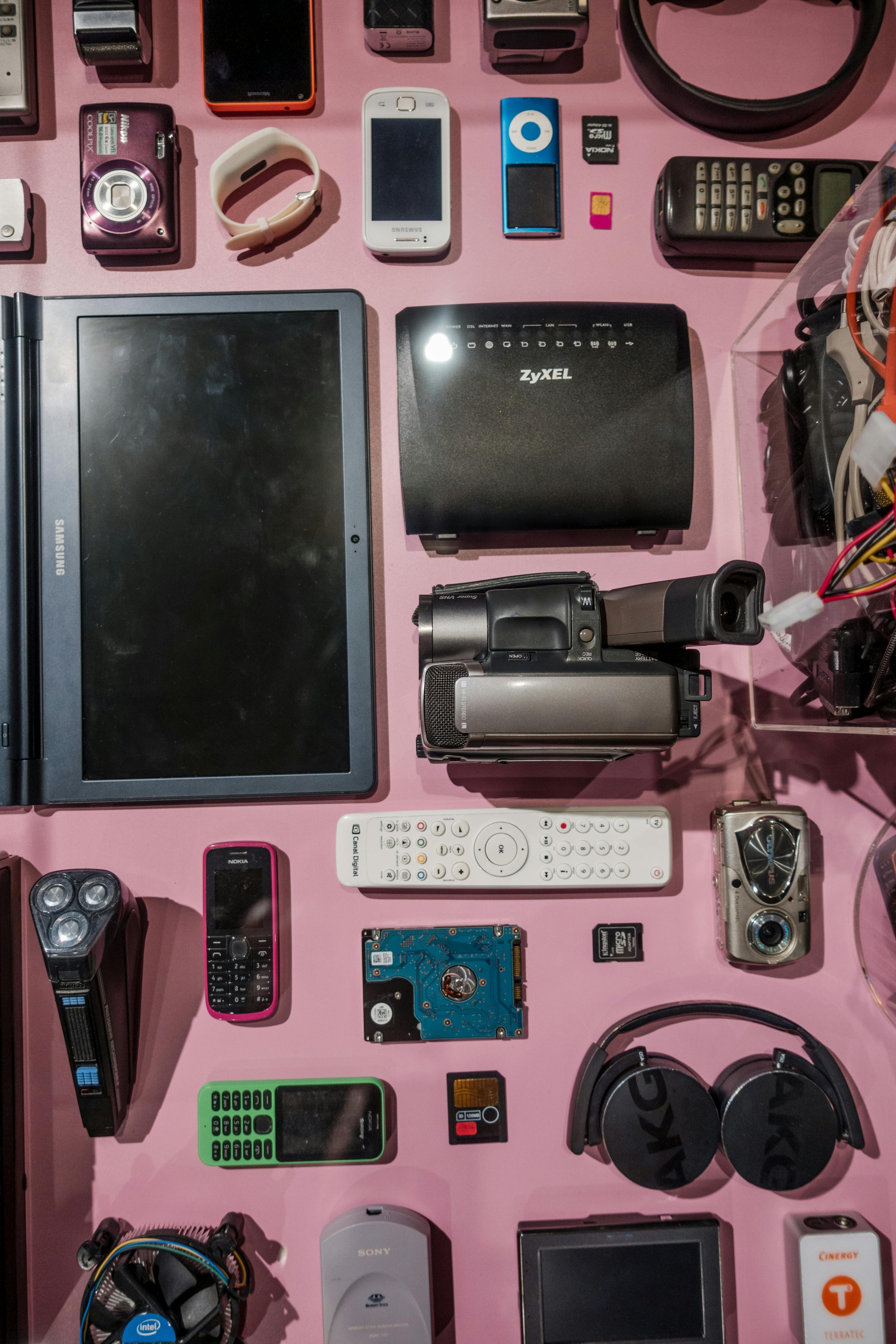 A collection of electronic devices on a pink surface, including cameras, mobile phones, a laptop, a Wi-Fi router, a power bank, a remote control, SD cards, computer hardware components, and headphones.