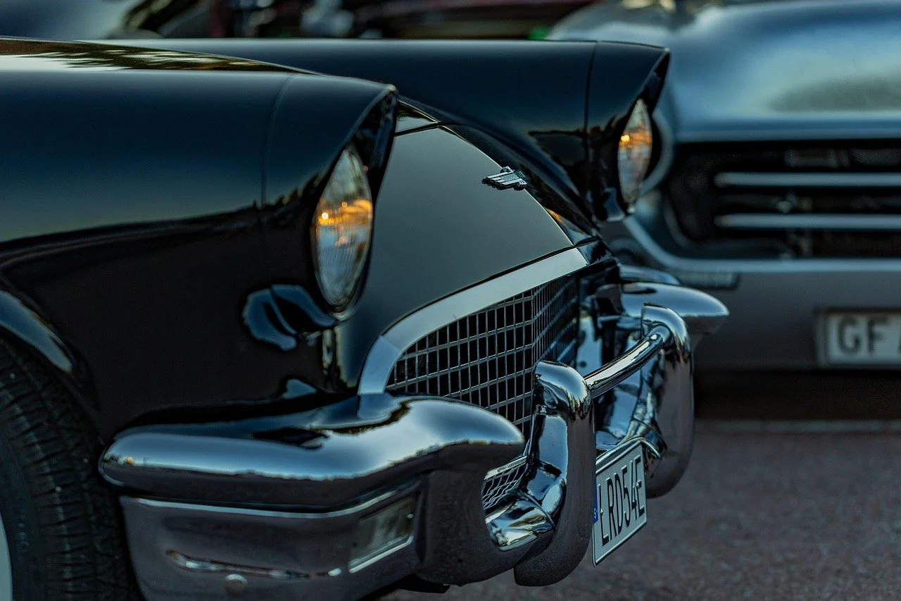 Close-up of the front ends of two vintage black cars parked side by side, showing their chrome bumpers, grills, and headlights in a parking lot during twilight.