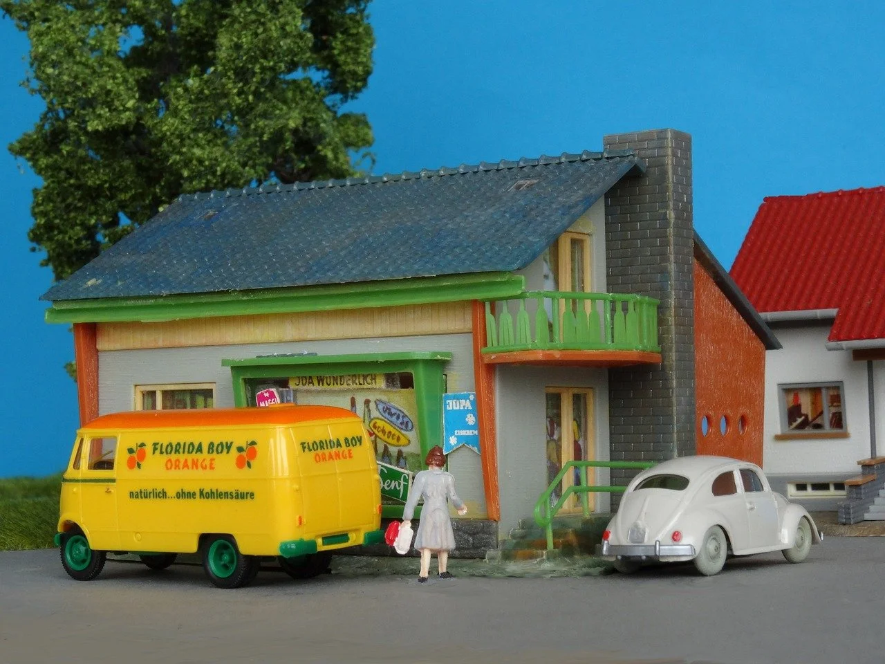 A miniature model scene of a street with a toy yellow van labeled "Florida Boy Orange," a small white toy car, and a tiny woman figurine holding a bag. The scene includes a detailed miniature house with a blue roof, green balcony, and brick chimney, set against a backdrop of trees and a blue sky.