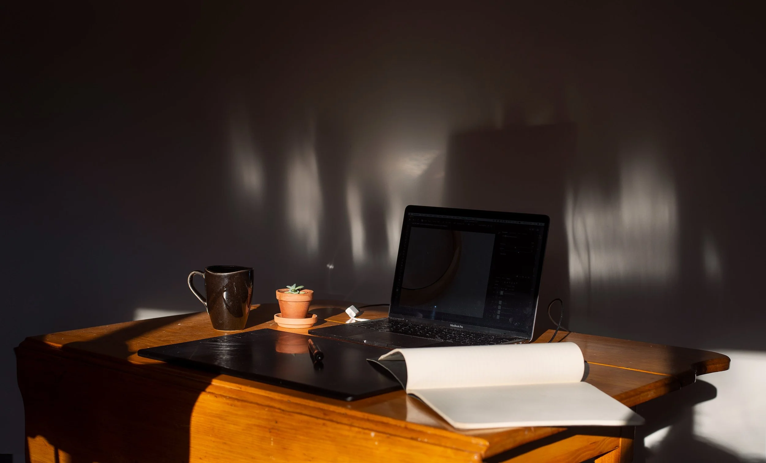A wooden desk with a MacBook Pro, an open notebook, a pen, a cactus in a small clay pot, a ceramic mug, and a black tablet or sketchpad. The background wall has shadows cast by light, creating shadows or patterns.