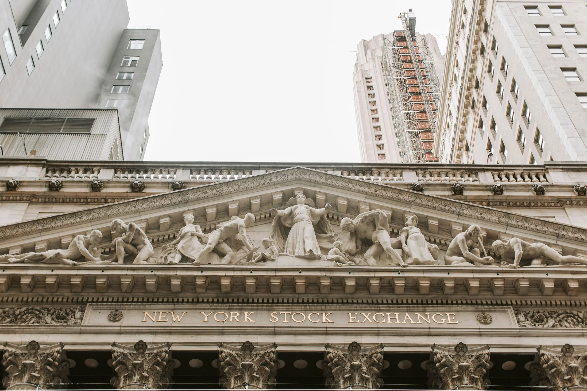 Facade of the New York Stock Exchange building with detailed marble sculptures and architecture, with tall buildings under a cloudy sky in the background.