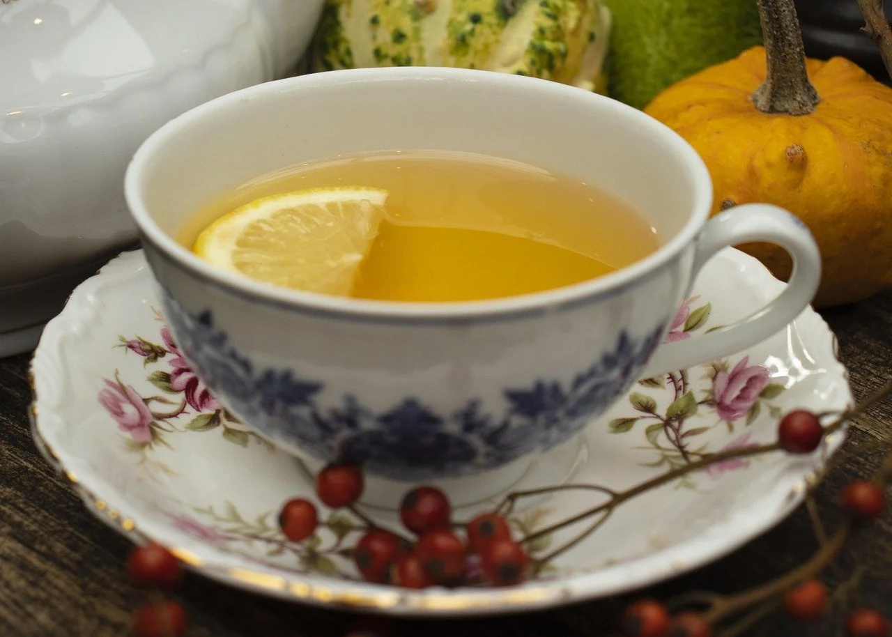 A cup of hot lemon tea with lemon slices in a floral patterned china cup, placed on a matching saucer, with berries in the foreground and pumpkins and gourds in the background.