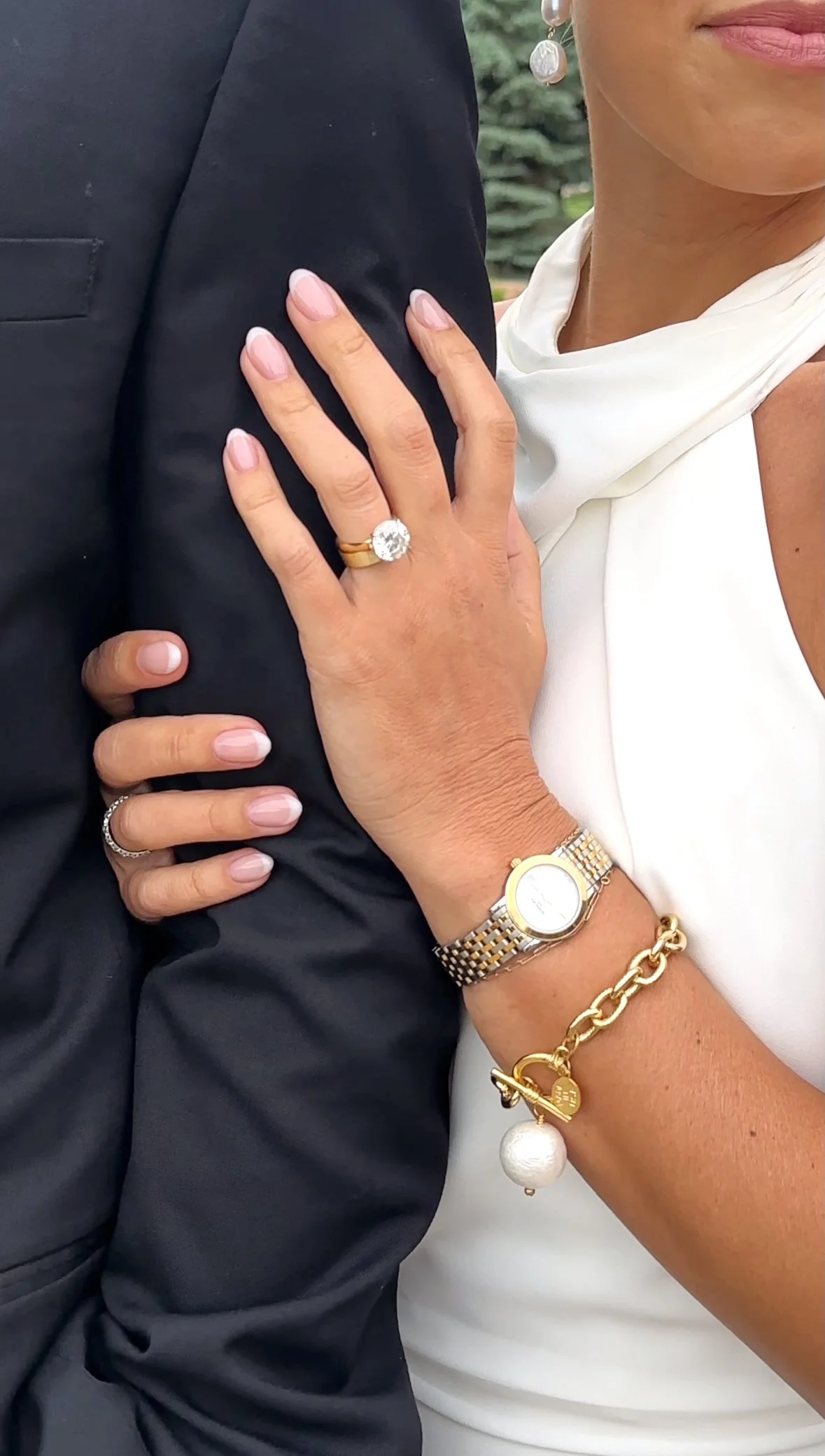 Close-up of a woman's hand with a large engagement ring resting on a man's shoulder. She is wearing a watch, gold bracelet, and a pearl charm bracelet. The woman is dressed in white, and the man in a dark suit.