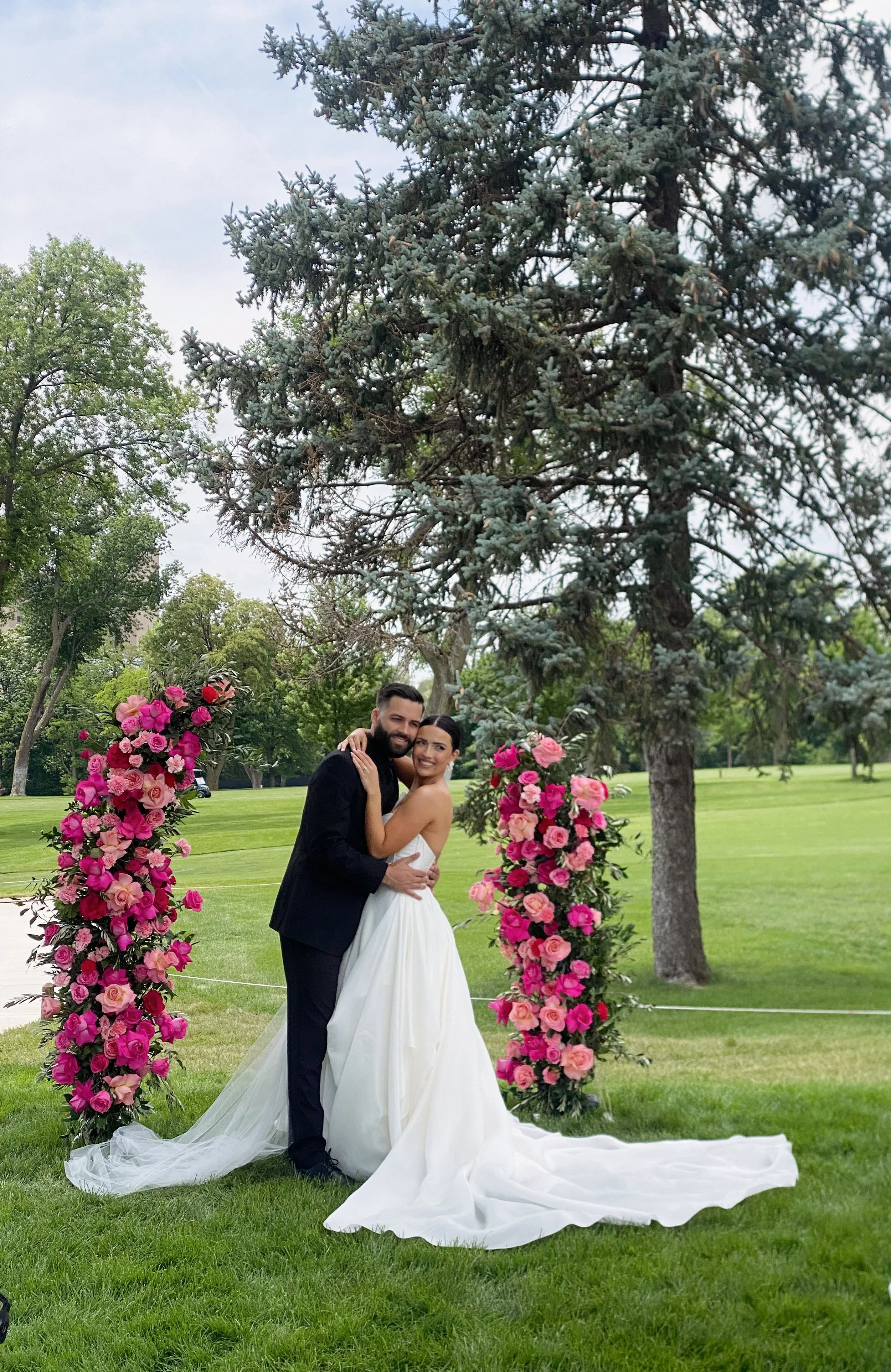 A newlywed couple embracing under a large pine tree, with floral arch of pink and purple flowers, in a lush green park.